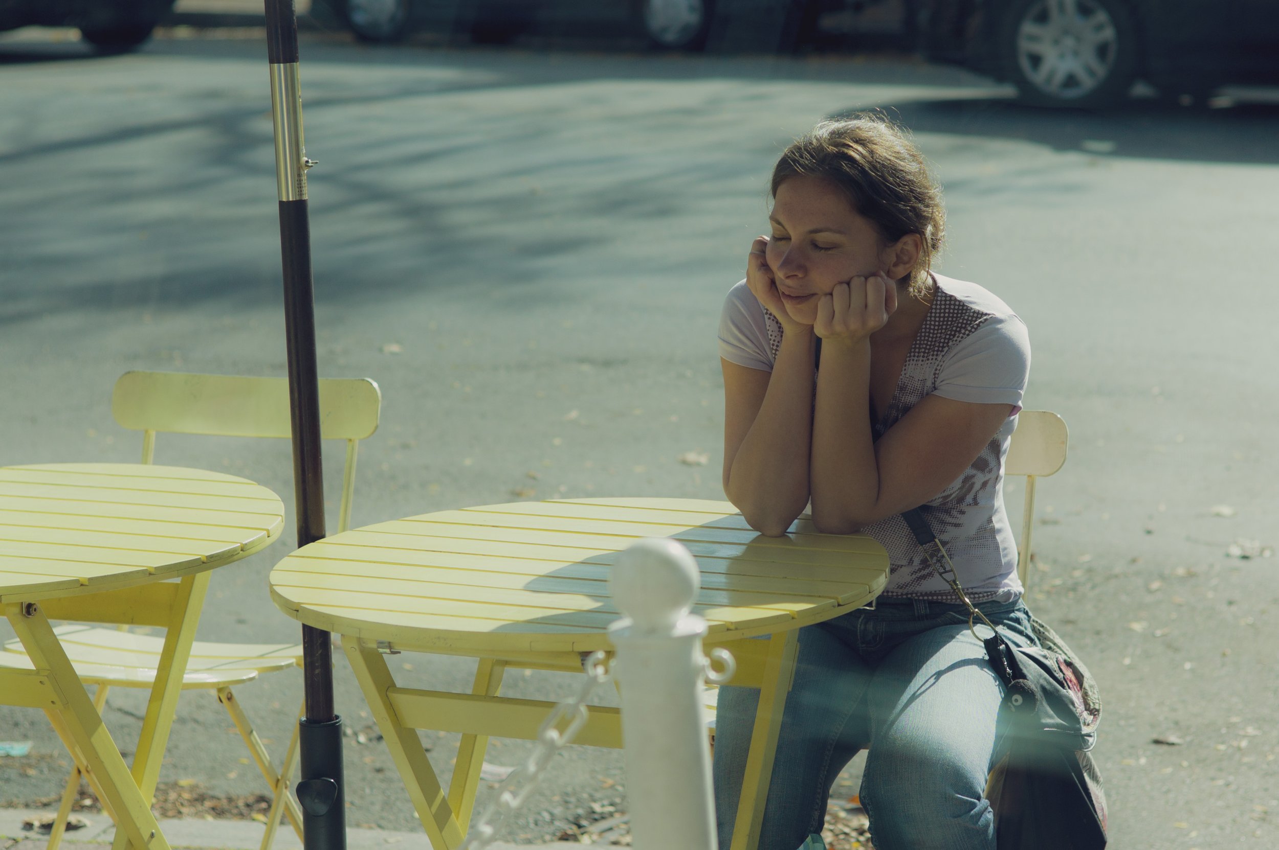 A woman crouching beside a yellow table with yellow chairs outdoors on a sunny day, resting her head on her hands and smiling gently.