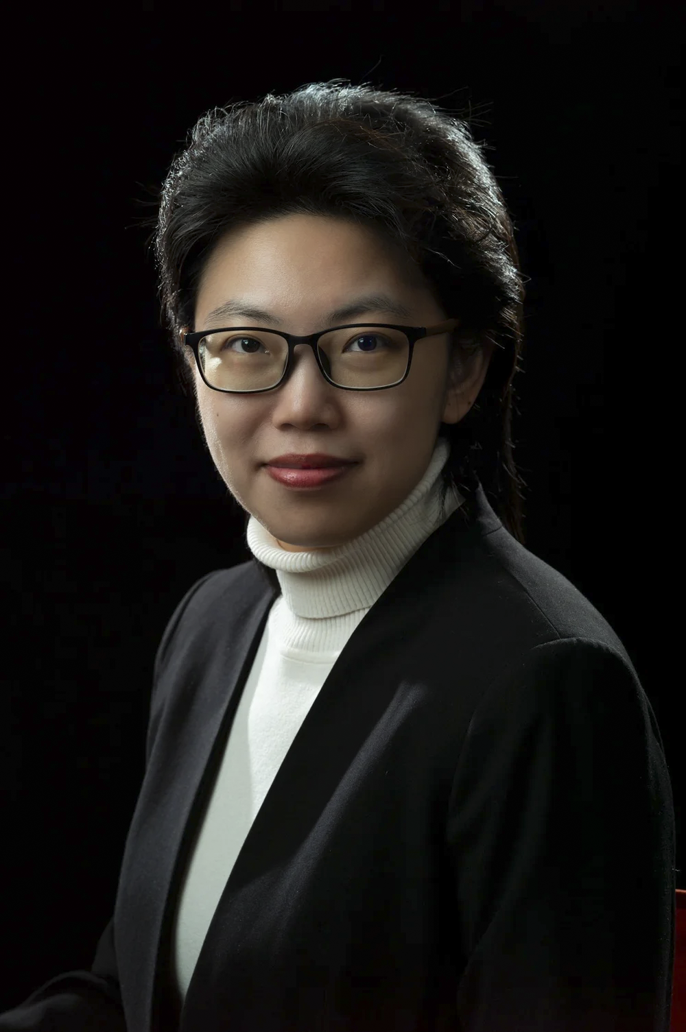 Professional portrait of a woman with glasses, wearing a black blazer and white turtleneck, against a dark background.