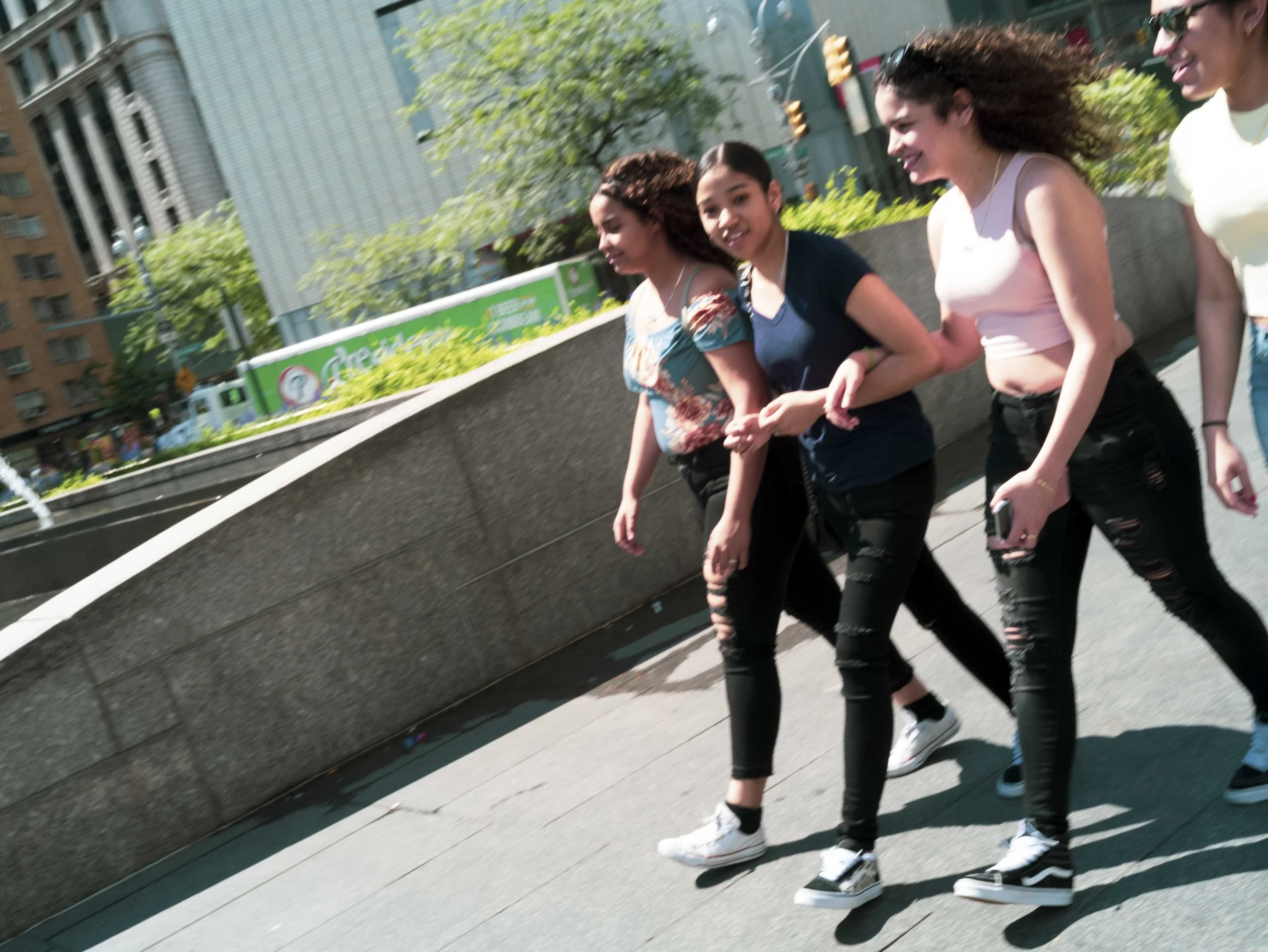 Four women walking together on a city sidewalk, smiling and enjoying the day. They are dressed casually, with two wearing sunglasses, and appear to be having a friendly conversation.