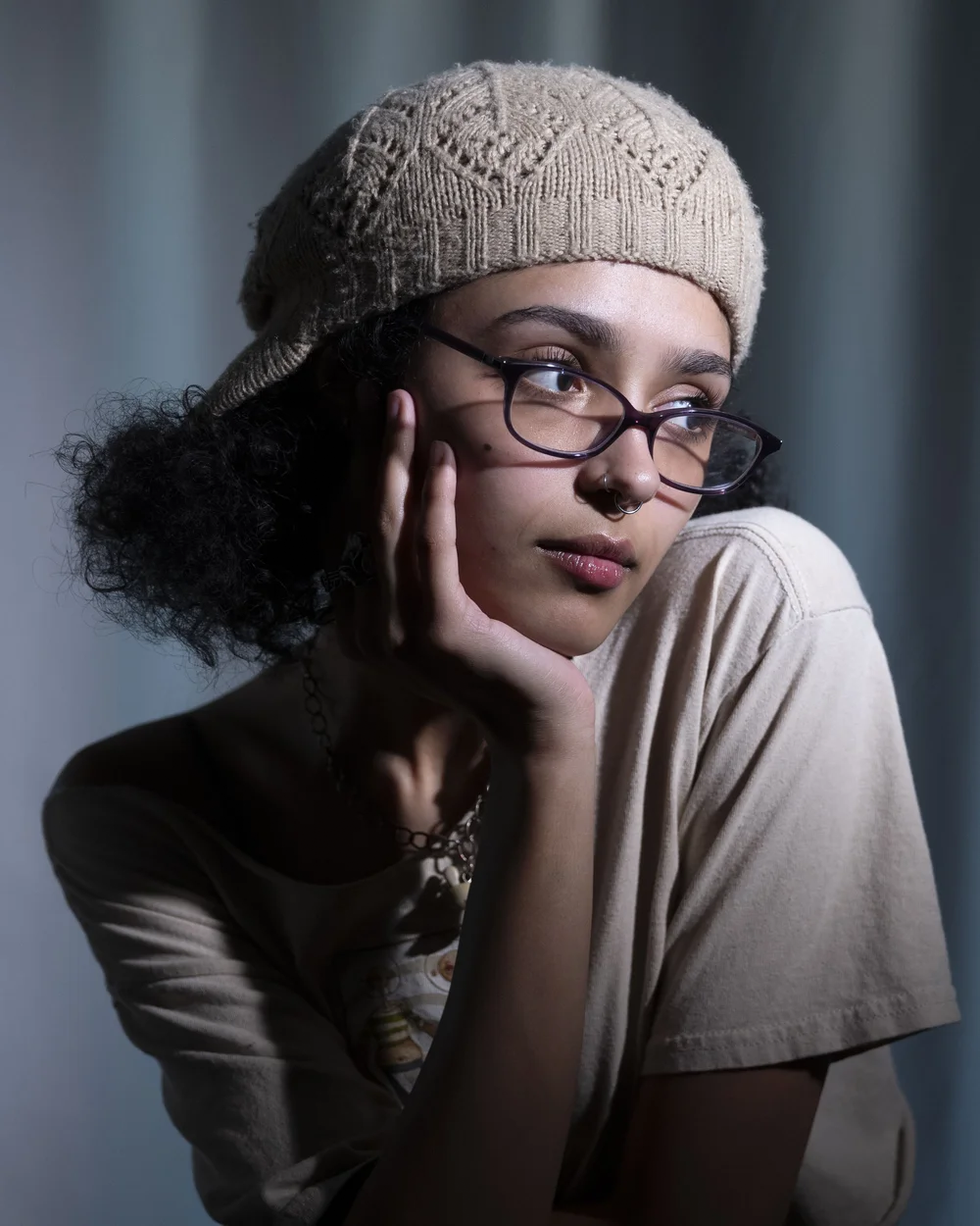 A woman with glasses and a septum piercing resting her chin on her hand, wearing a beige beanie and a beige t-shirt.