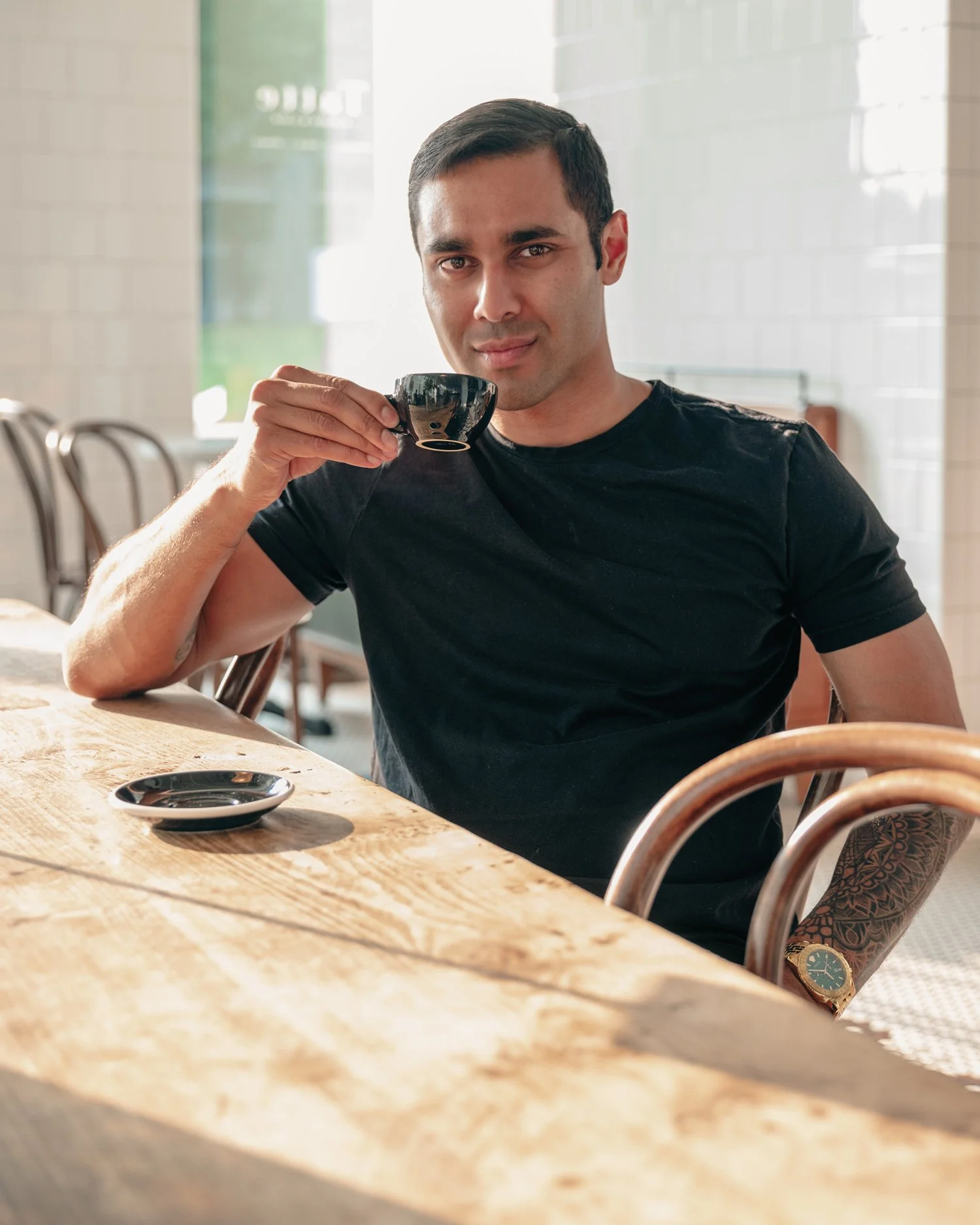 A man with short dark hair and a tattooed arm is sitting at a wooden table in a café, holding a black cup and looking at the camera, with a blurred background of chairs and windows.