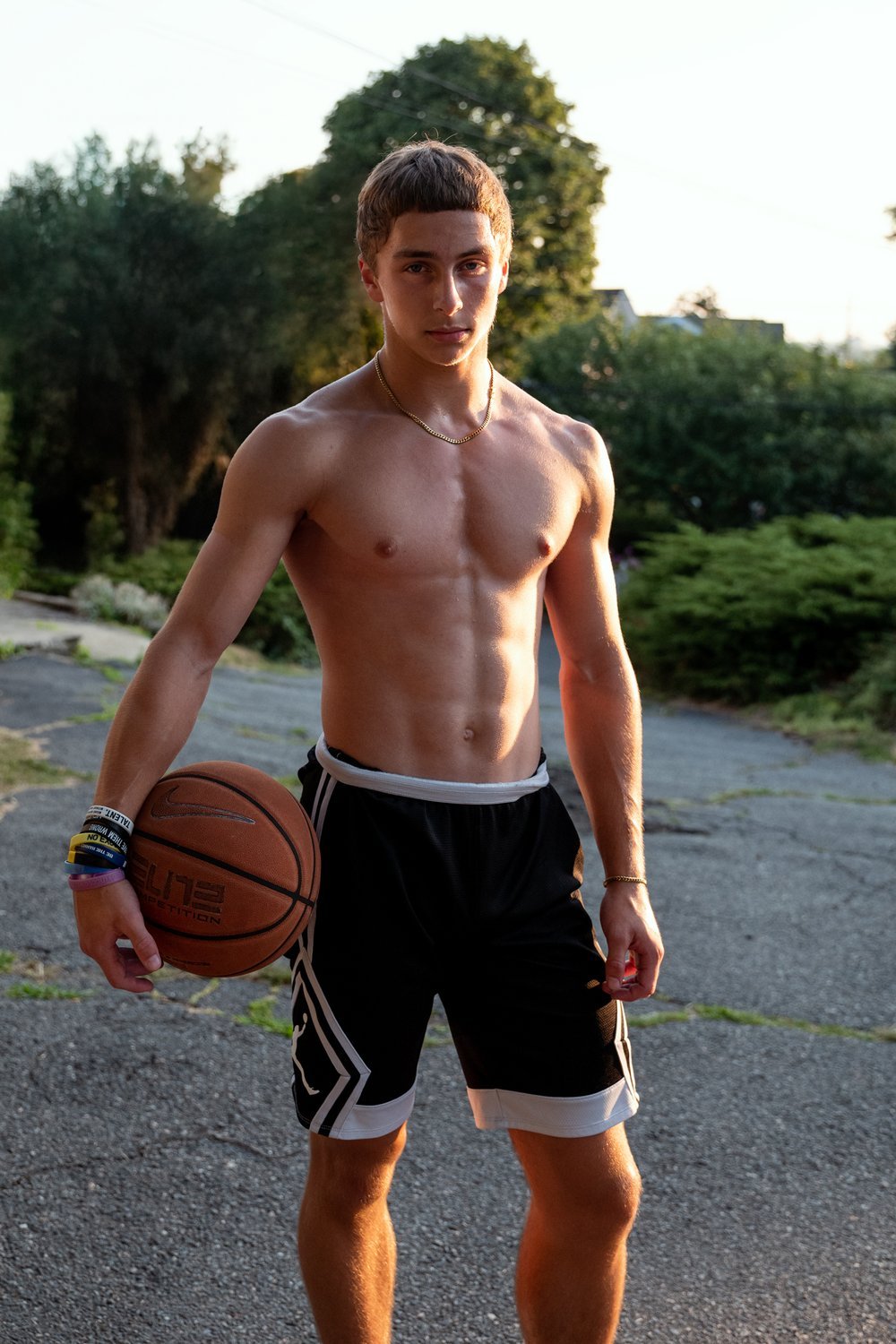 A shirtless young man holding a basketball outdoors during sunset, wearing black basketball shorts with white accents and multiple wristbands on his left wrist, with trees and houses in the background.
