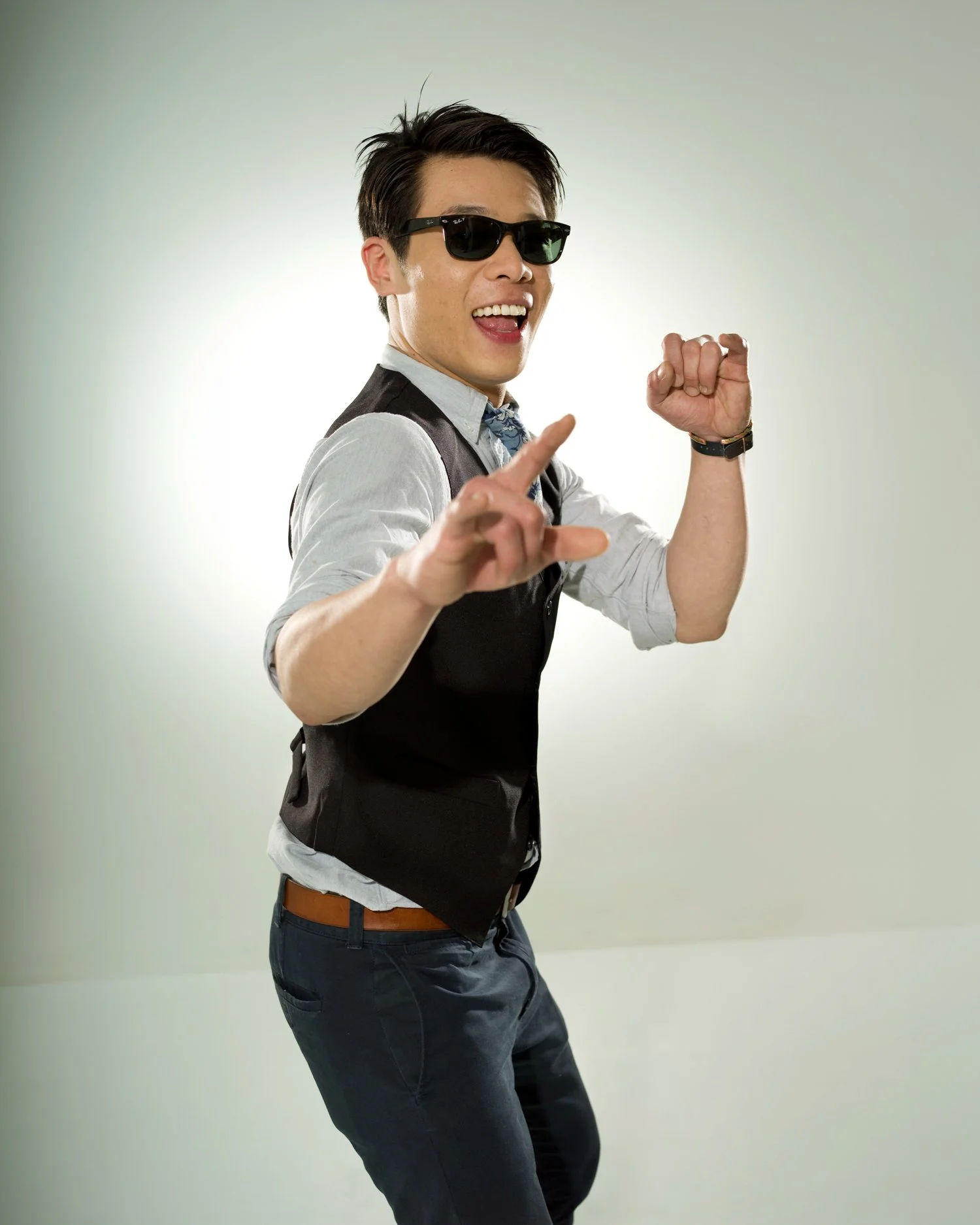 A young man wearing sunglasses, a white shirt, a black vest, and jeans, smiling and pointing upward with one hand and making a fist with the other, against a plain light background.