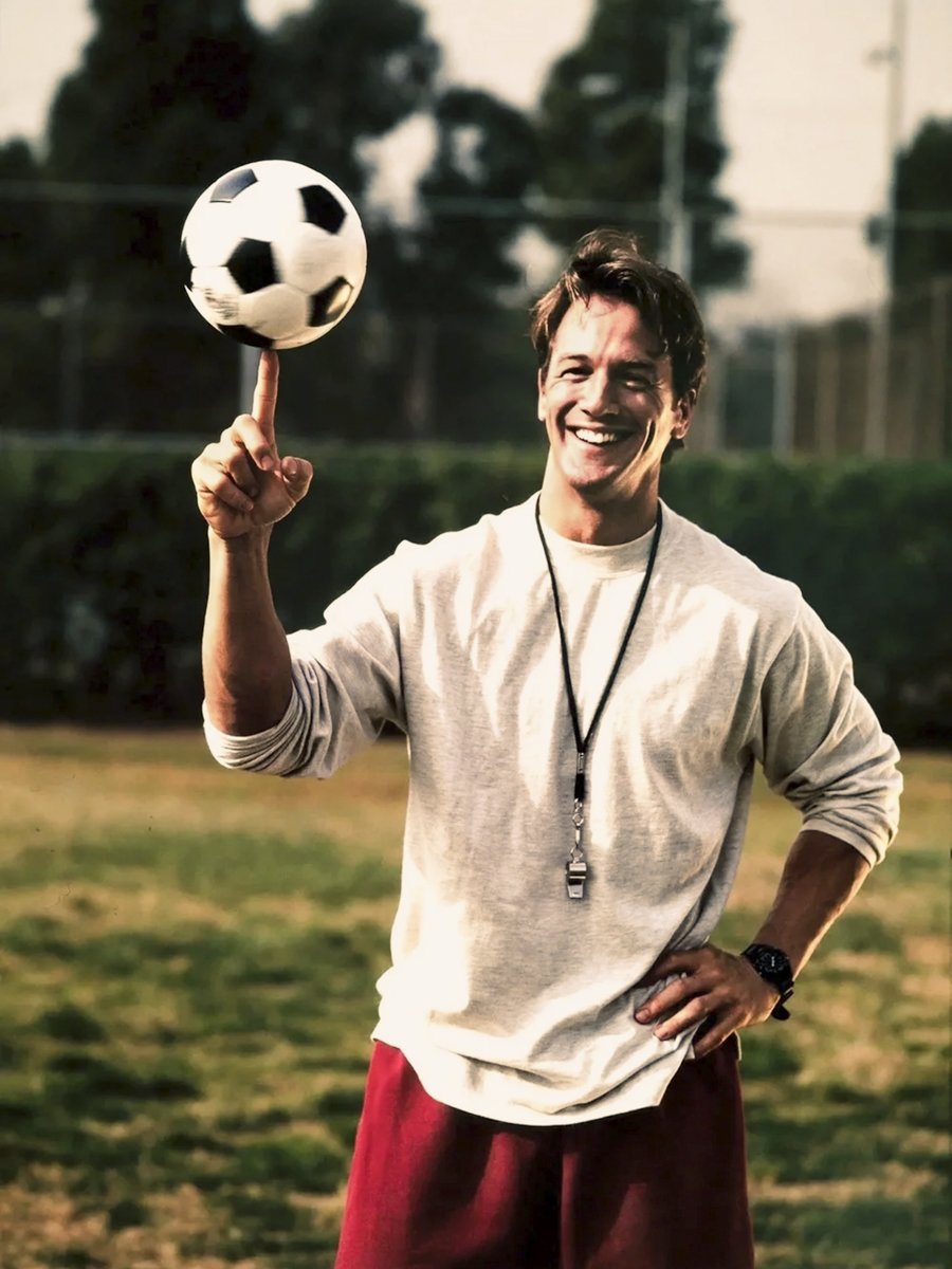 Young male coach spinning a soccer ball on his forefinger in an outdoor park setting with late afternoon golden light.
