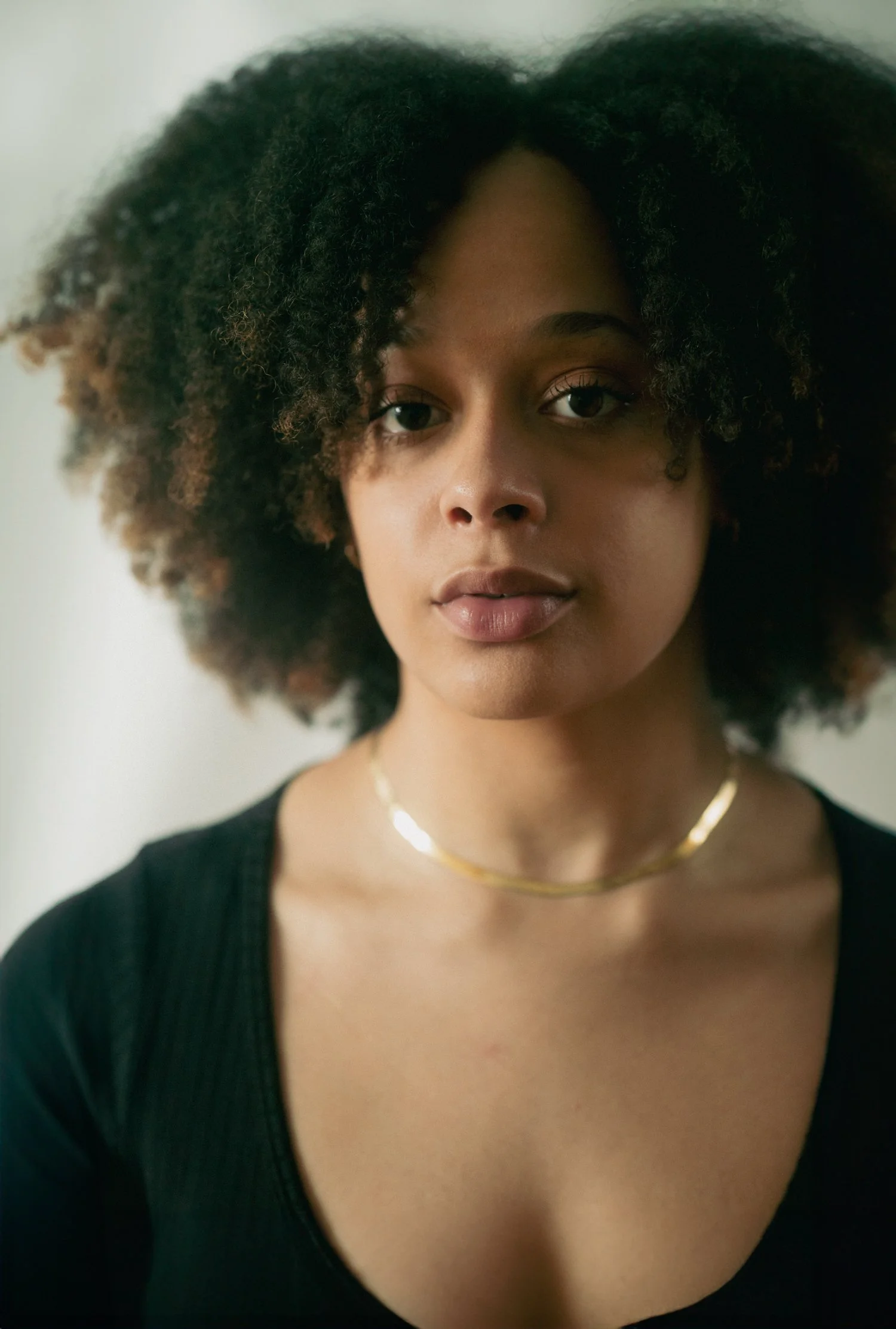 A young woman with curly black hair, wearing a black top and a gold necklace, poses in natural light against a plain background.