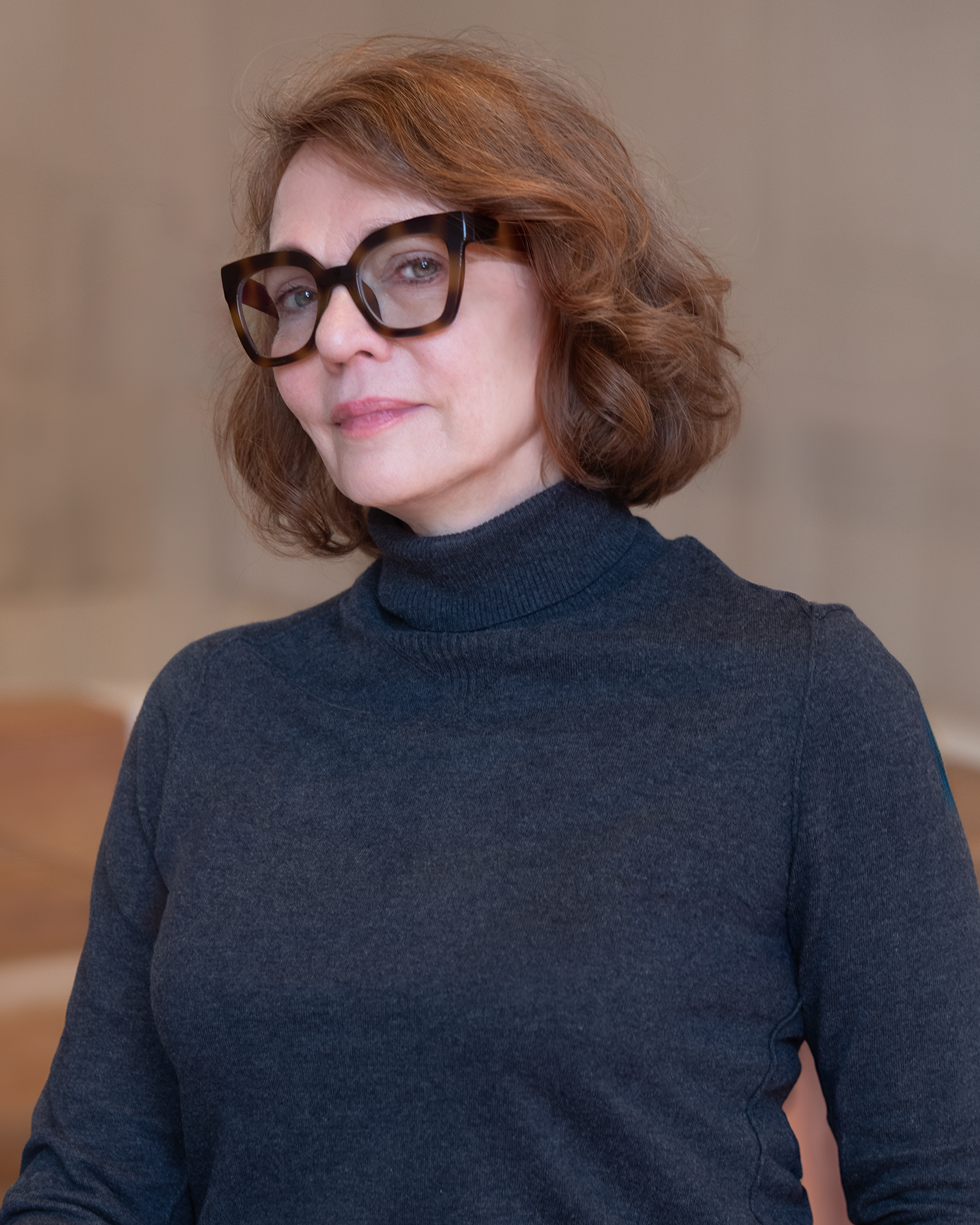 A woman with copper-red hair wearing thick-rim, tortoise shell glass with a dark turtleneck top. She is seen from waist up with her head in a three-quarter view as she makes eye-contact with the viewer. Photographed at an indoor location with a mediu