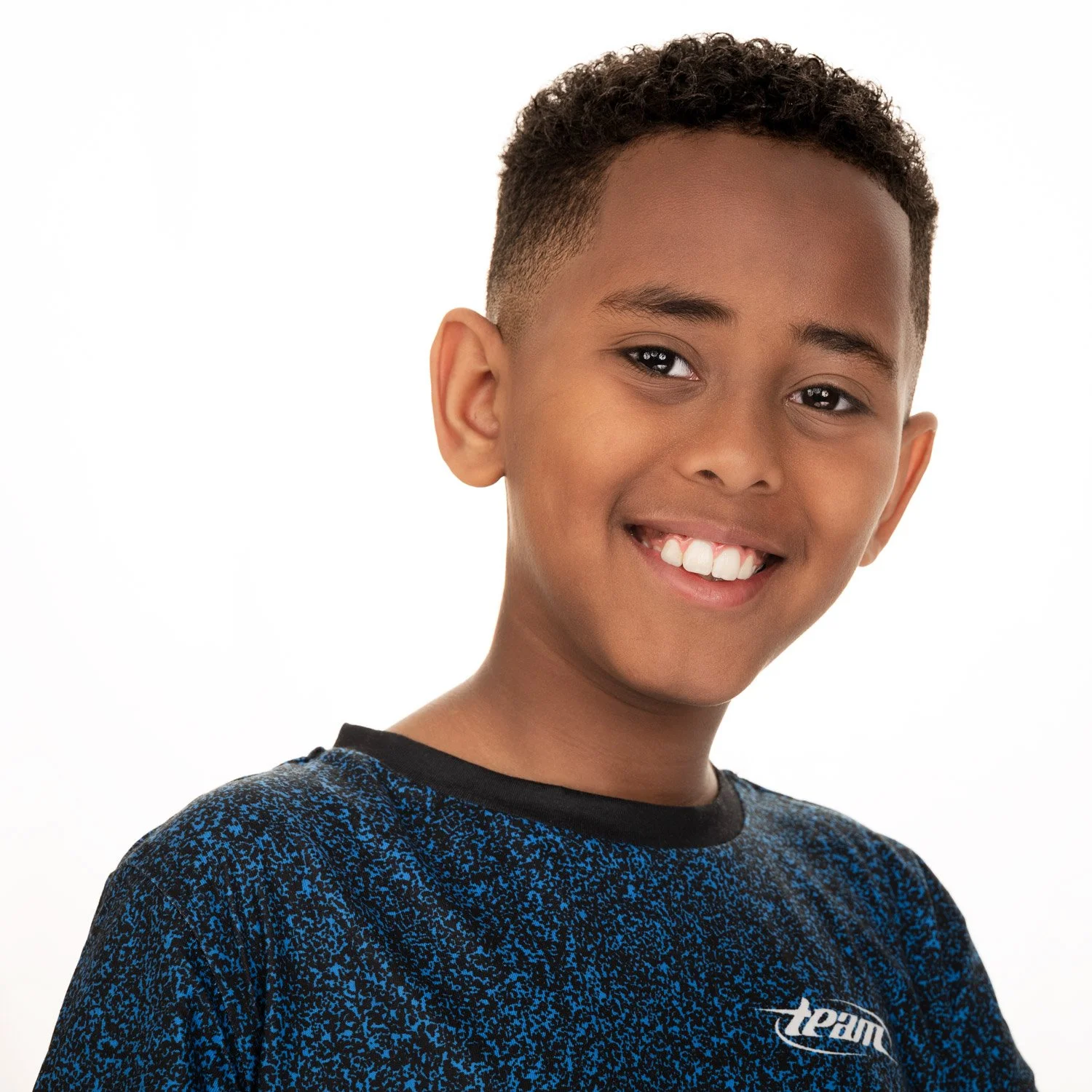 A young boy with short curly hair and a big smile showing his teeth, wearing a dark blue and black patterned shirt with the word 'dream' on it, against a plain white background.