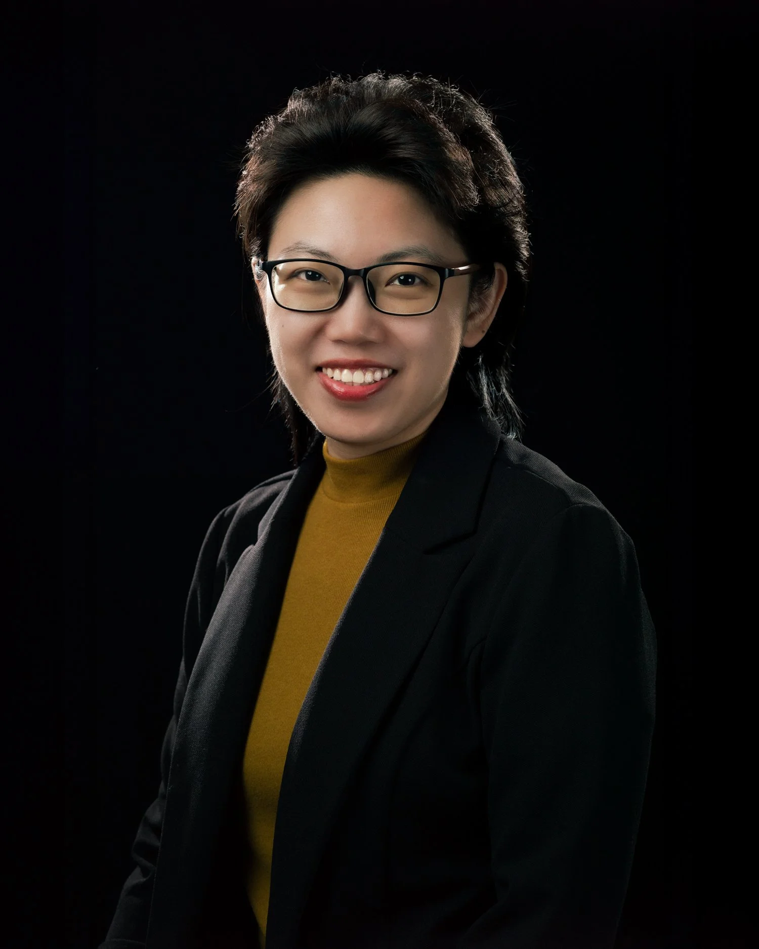 Professional portrait of a woman with glasses, wearing a black blazer and gold turtleneck, against a dark background.