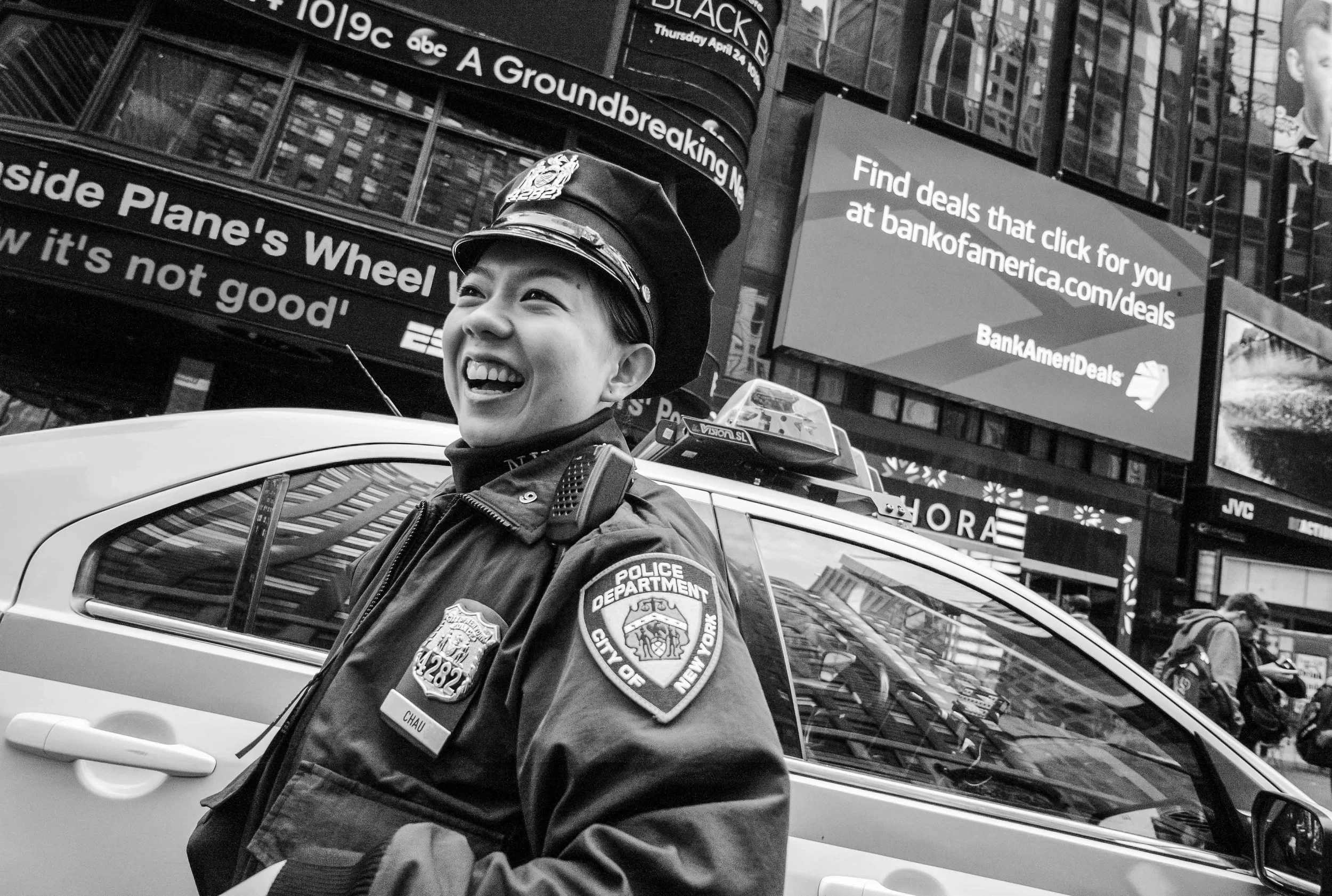 A black and white photo of a smiling female police officer in uniform standing next to a vehicle in an urban setting with bright electronic billboards and buildings in the background.