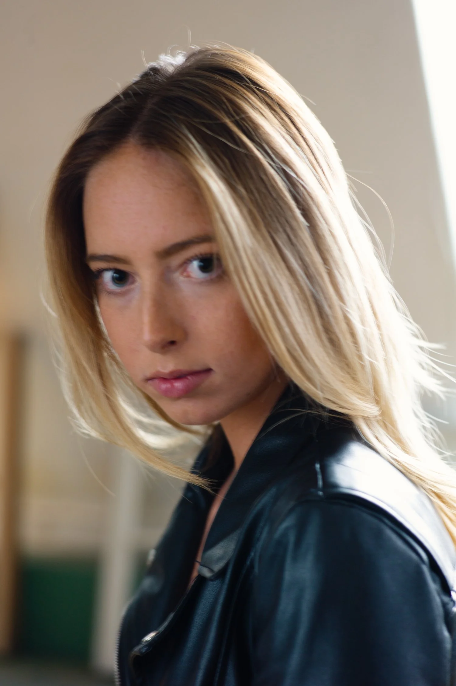 Close-up of a young woman with blonde hair and blue eyes, wearing a black leather jacket, indoors.