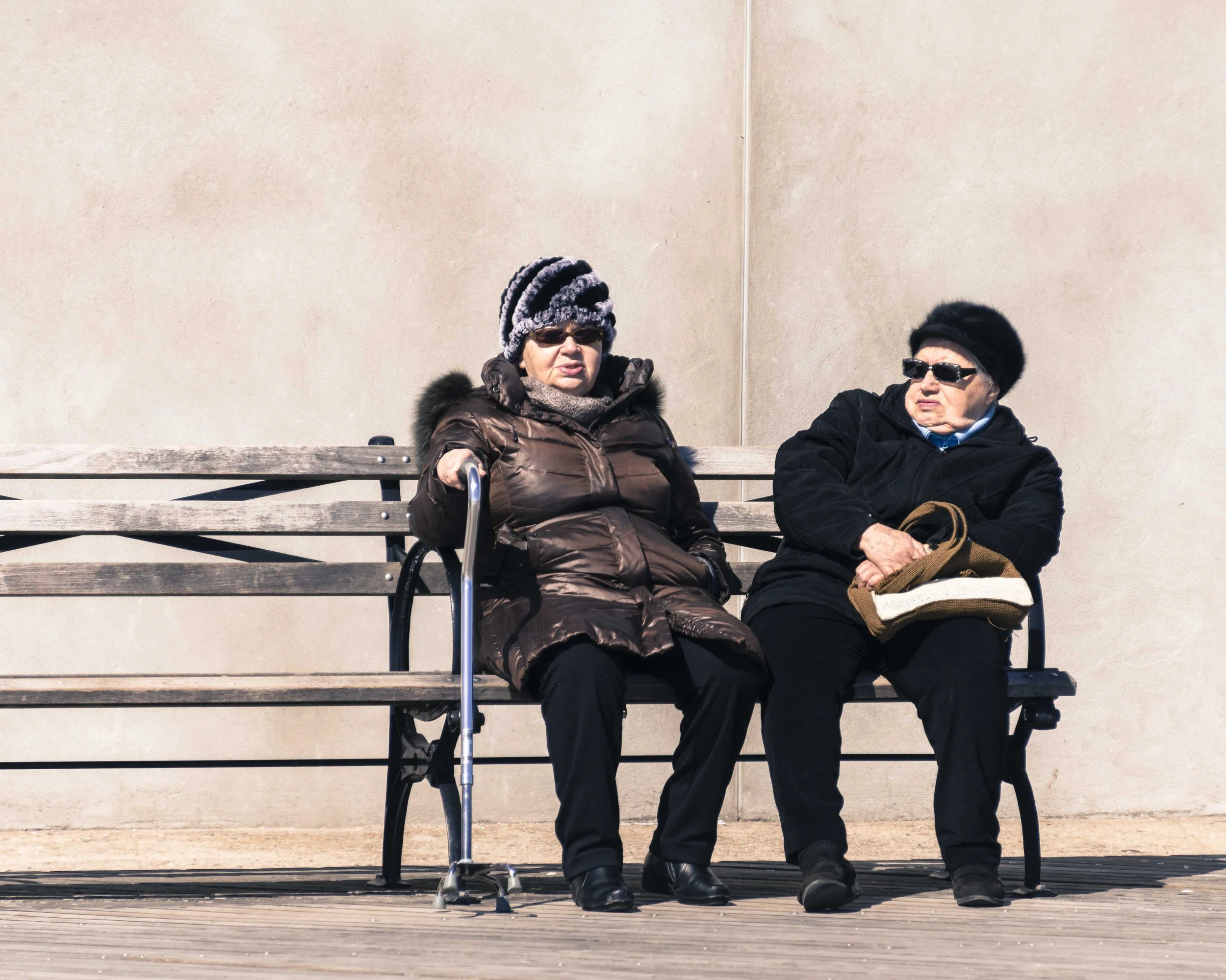 Two elderly women wearing winter coats and hats sitting on a wooden bench against a beige wall. One woman is wearing sunglasses and holding a cane, while the other has dark glasses and a hat.