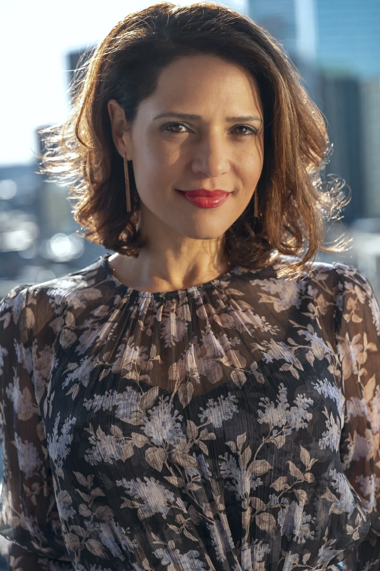Actress Monique Gabriella Curnen in a natural light portrait shot outdoors in sunlight against a city background. She has shoulder-length brown hair and red lips and wear a dress with sheer fabric in brown and white floral print.