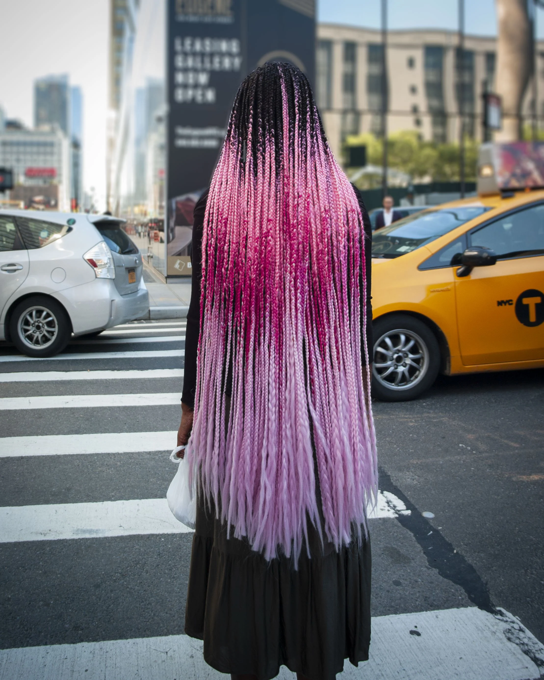Person with long pink and black braided hair standing at a crosswalk in an urban setting, facing away from the camera, with cars and a yellow taxi around.