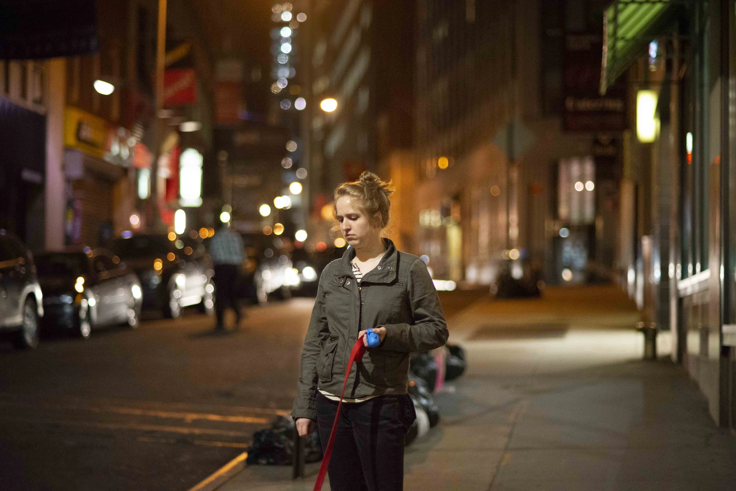 A young woman with blonde hair tied up in a bun walking on a city street at night. She is wearing a gray jacket and looking down at something in her hand. The street is lit with streetlights, and there are parked cars and storefronts along the sidewa