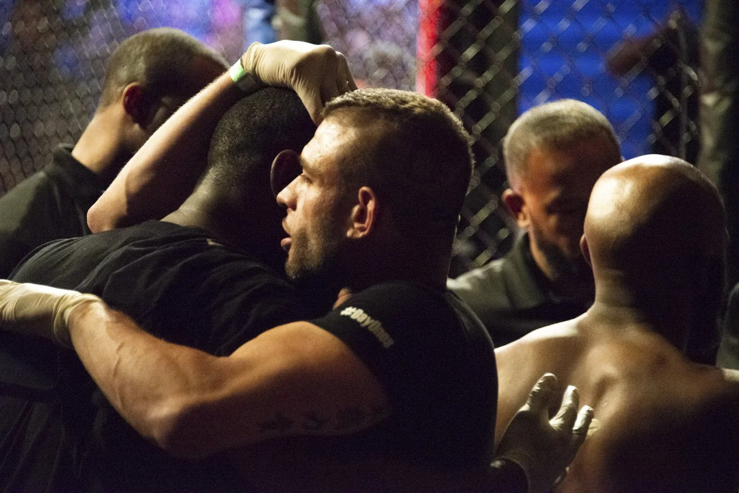 Mixed martial arts fighters and trainers inside an octagon cage, embracing and comforting each other after a match.