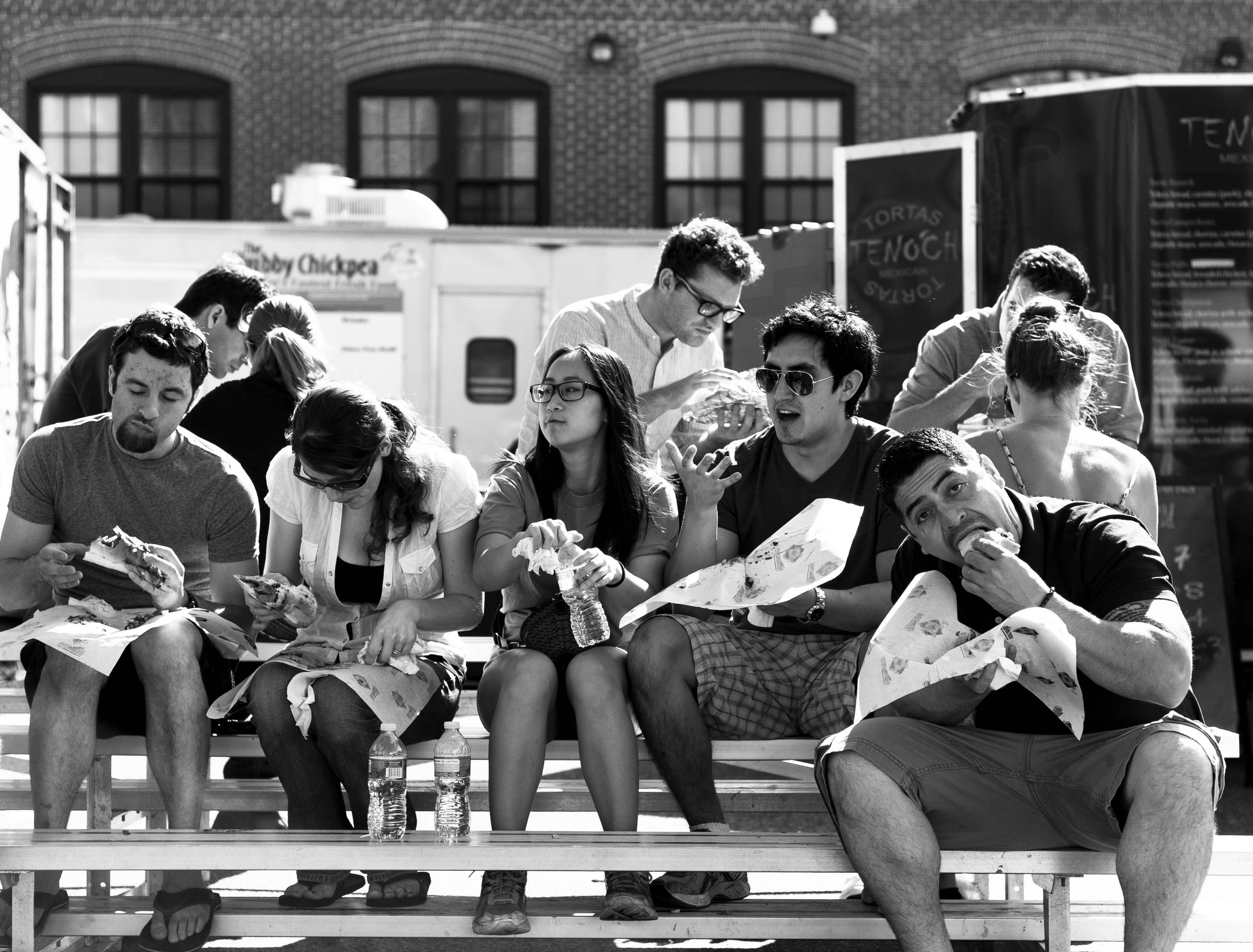 Group of young people sitting on stadium bleachers outside, eating food and drinking water bottles, with two food trucks and brick building in background.