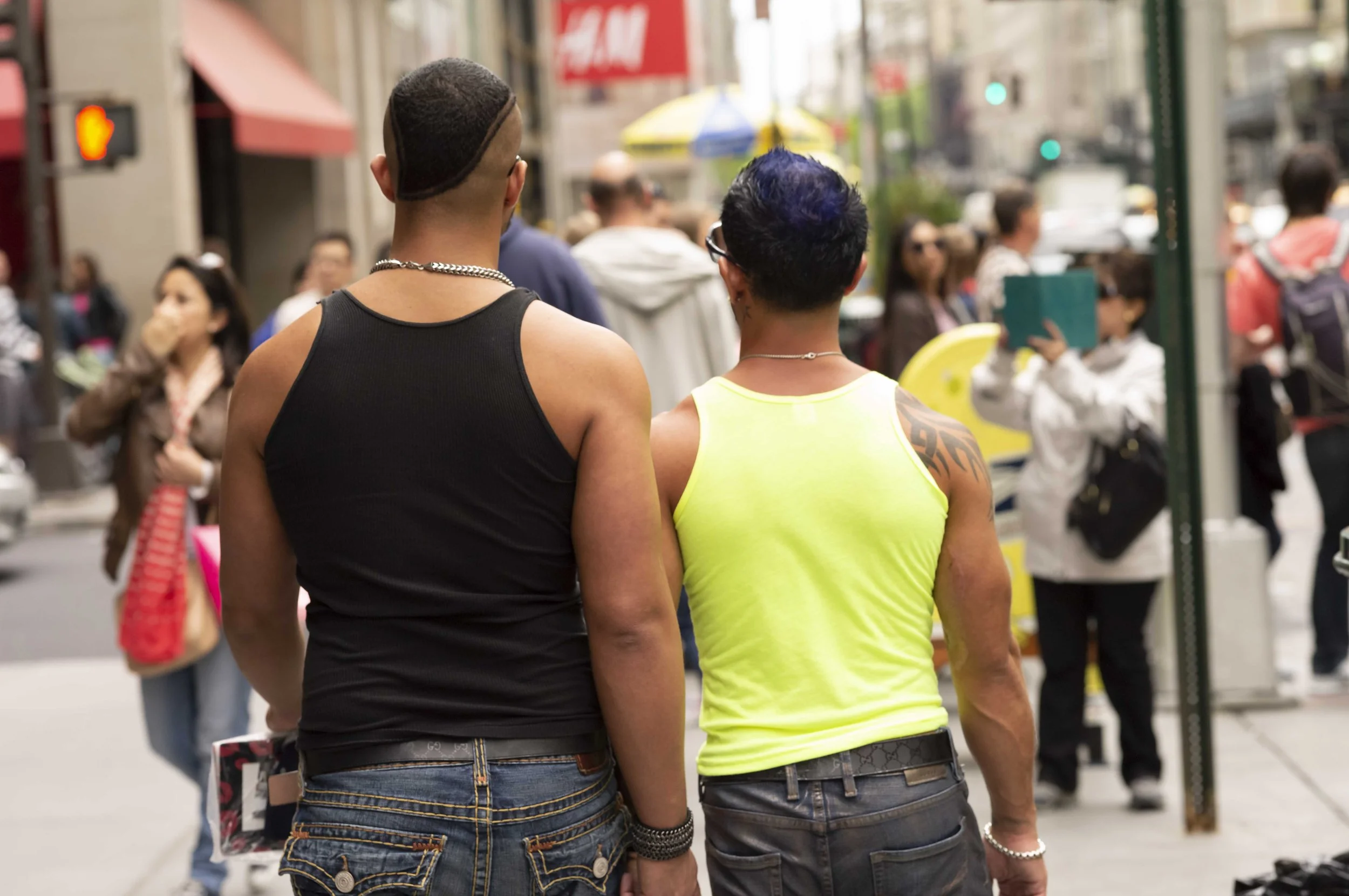 Two people standing side by side in a busy city street with many pedestrians and storefronts, viewed from behind. The person on the left has a mohawk hairstyle and wears a black tank top. The person on the right has short dark hair with purple highli