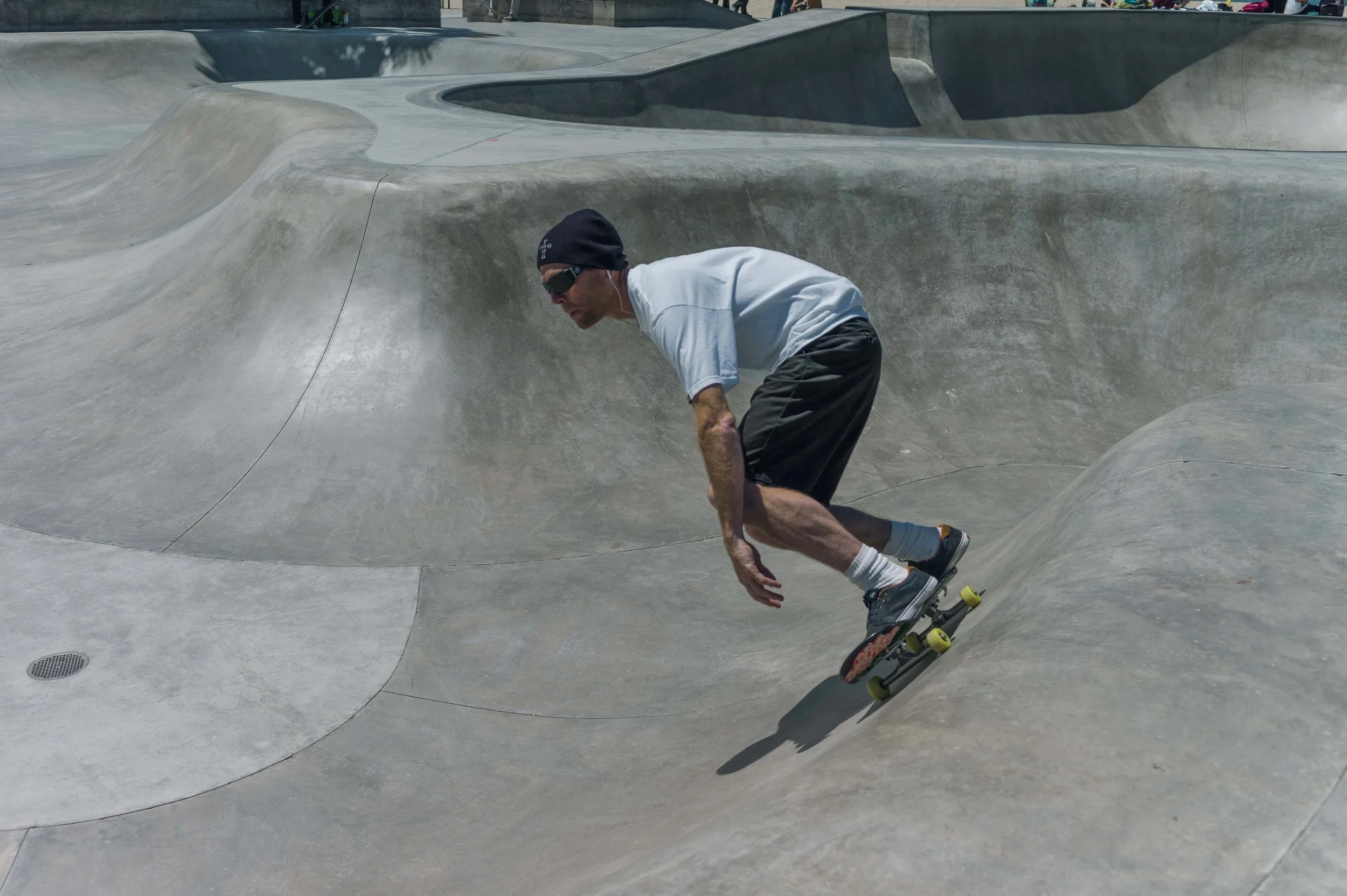A man in a white T-shirt, black shorts, a black beanie, and sunglasses skating at a concrete skatepark with smooth, curving surfaces.