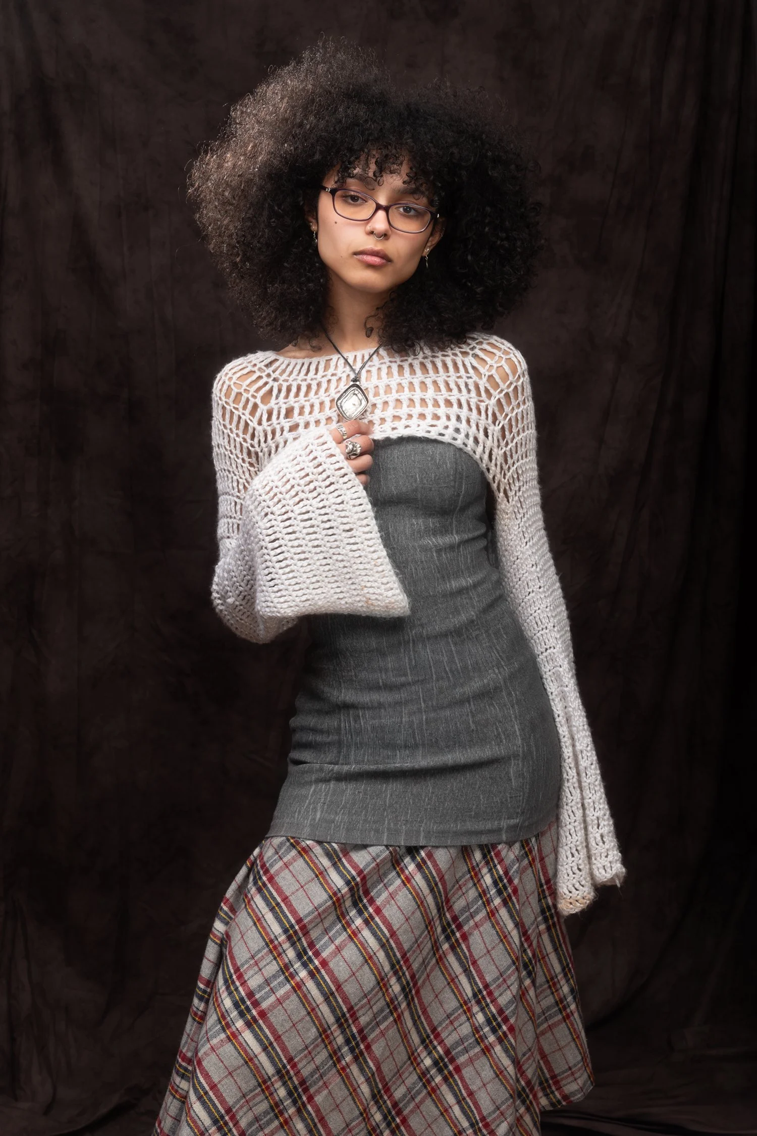 Portrait of a young ethnic woman with black, tightly-curled hair and heavy-rimmed glasses posing in front of a dark fabric studio backdrop. She wears a white knit, homemade crop-top with bell sleeves over a snug, gray mini dress over a long plaid ski