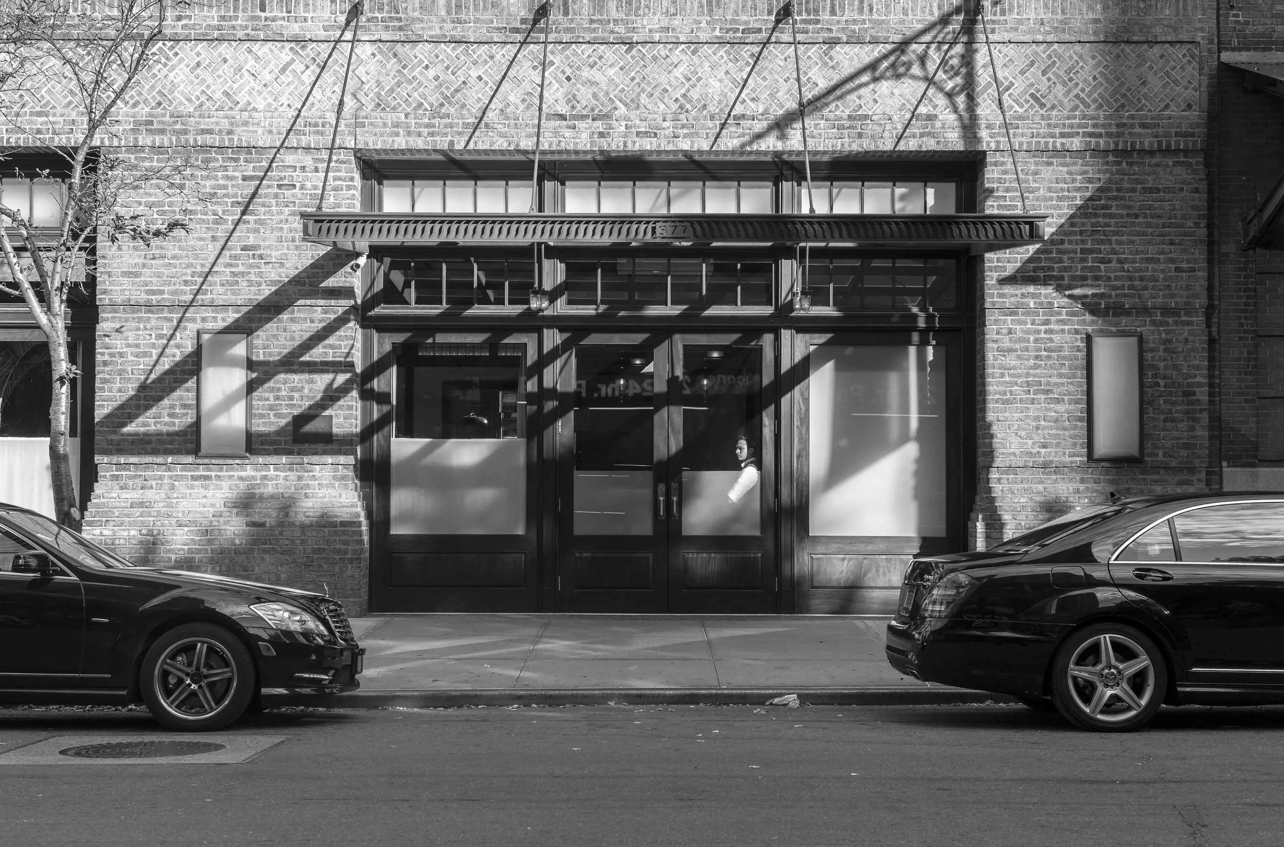 Black and white photo of a brick building with a large windowed door. Two cars are parked in front of the building on the street. A person is seated inside the door, looking to the side.