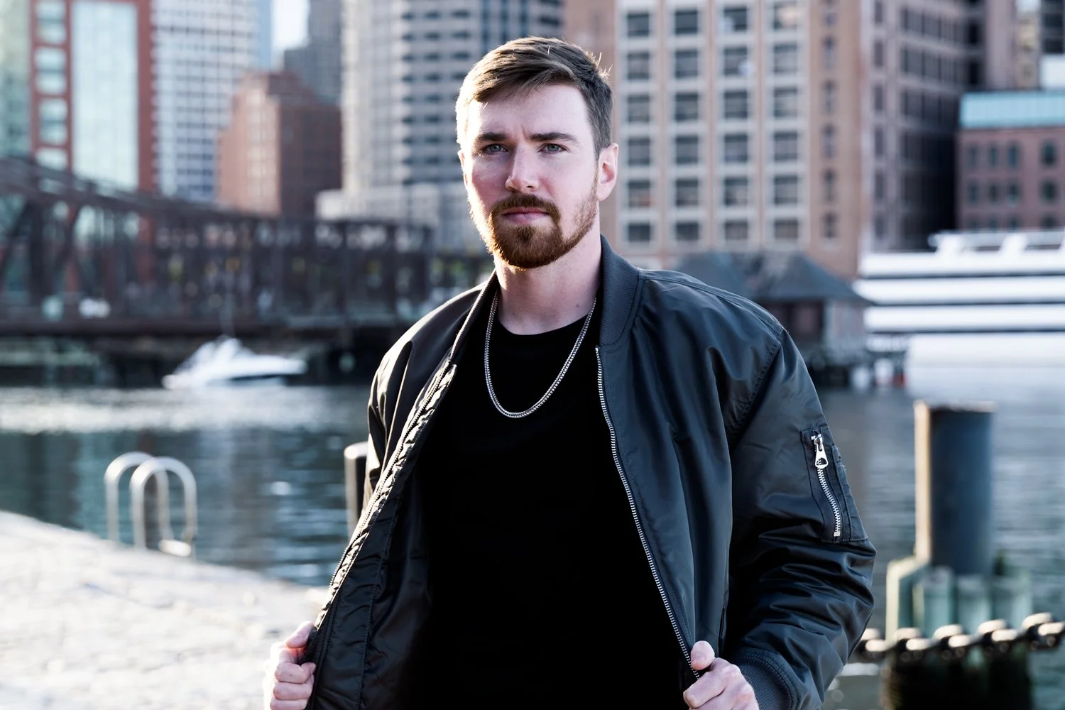 A young man in a bomber jacket and black t0shirt wears a silver neckchain while standing at the waterfront in the South Boston Seaport district.