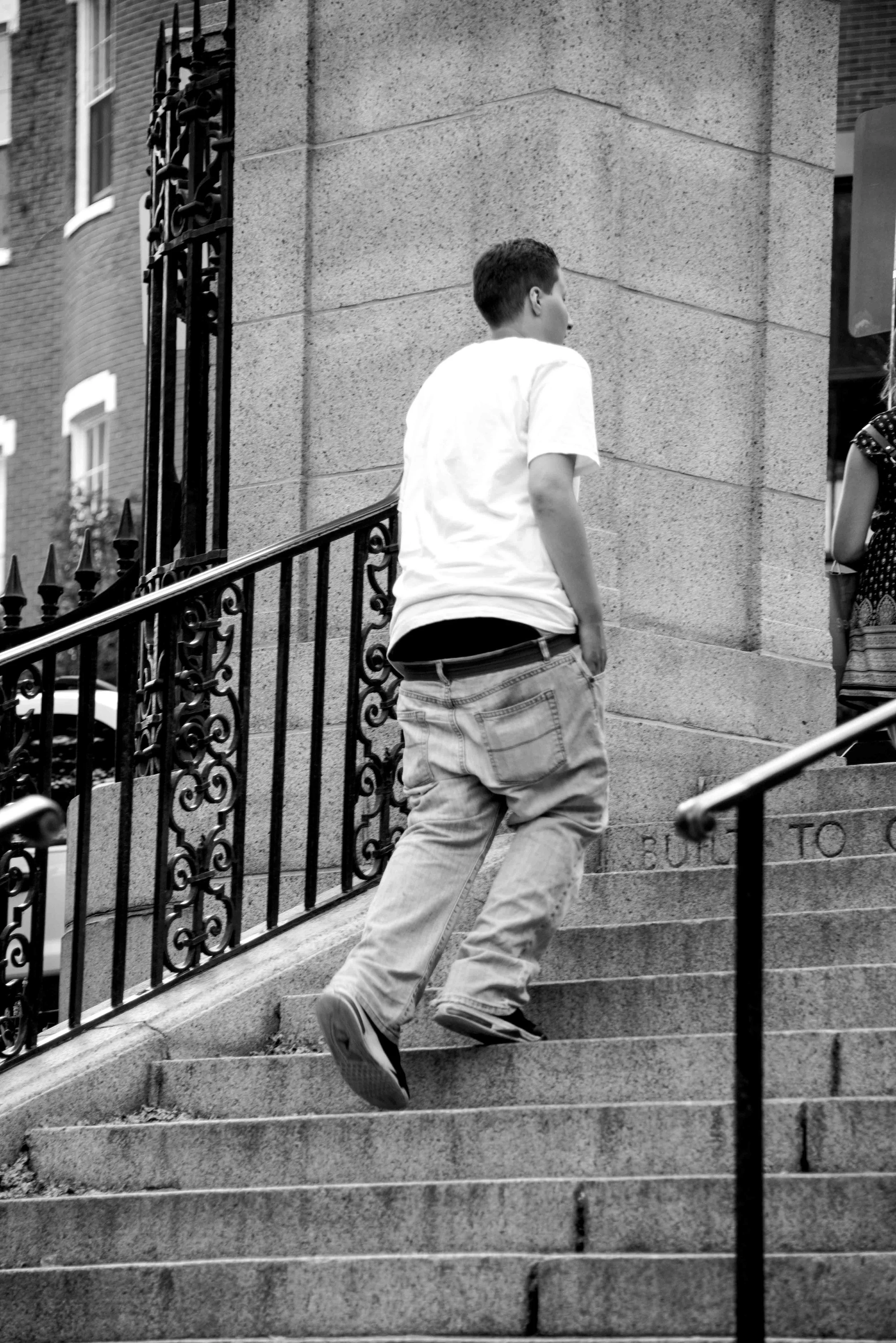 A young person in a white t-shirt and baggy jeans is walking up outdoor stone stairs with a railing, near a brick building, with part of another person visible on the right.