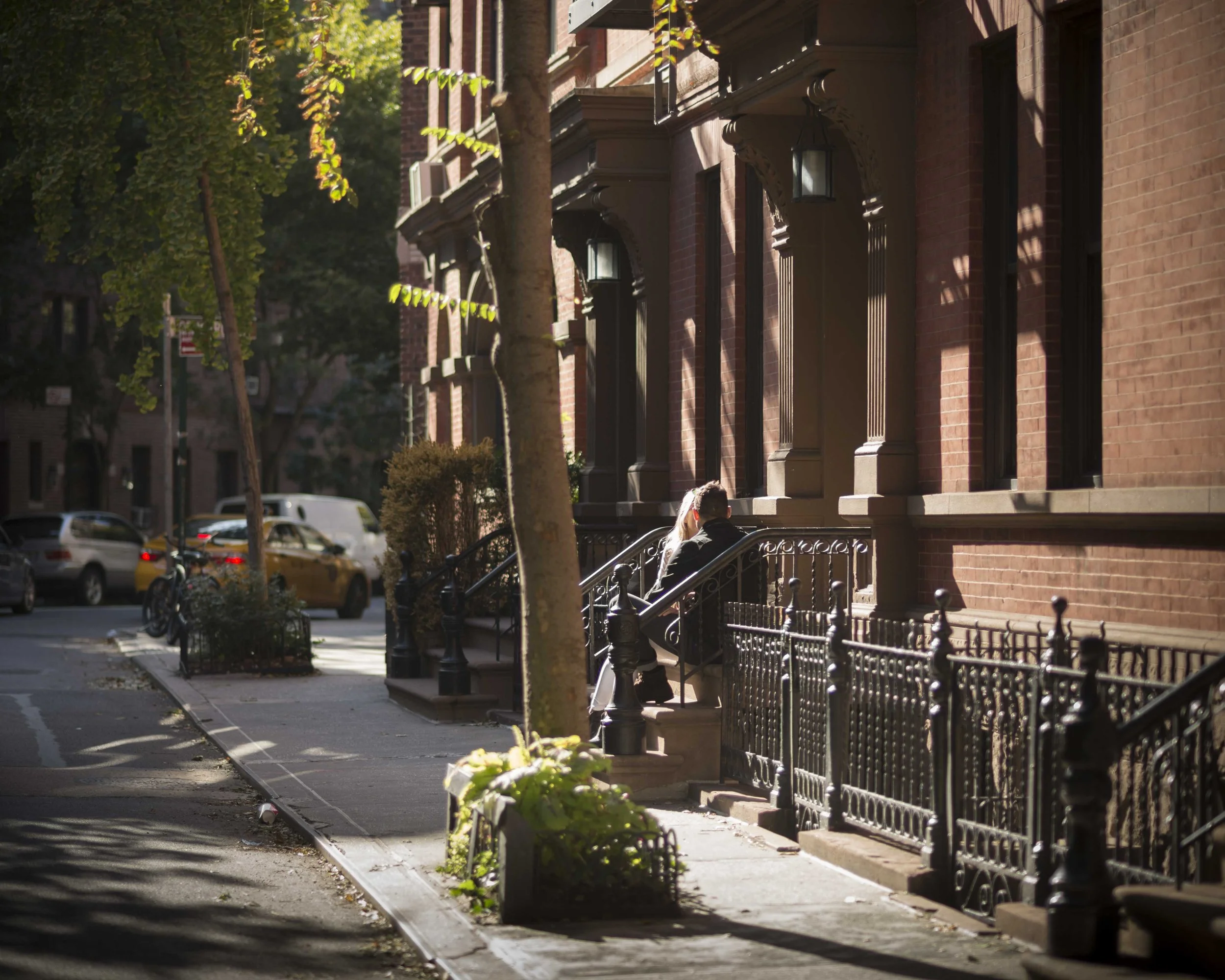 A city sidewalk with residential buildings, parked cars, trees, and a person sitting on stairs outside a red brick building.