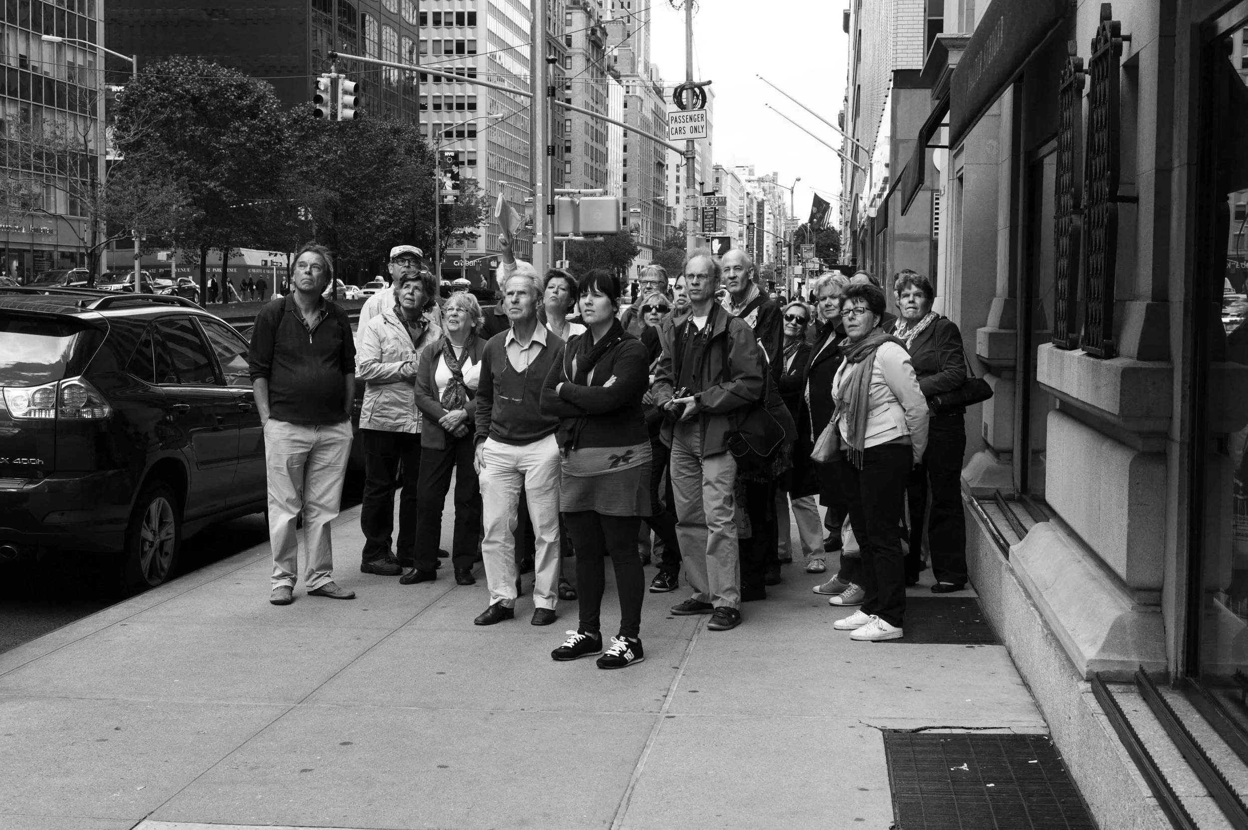 A group of people standing on a city sidewalk, looking up, with tall buildings and traffic lights in the background, in black and white.