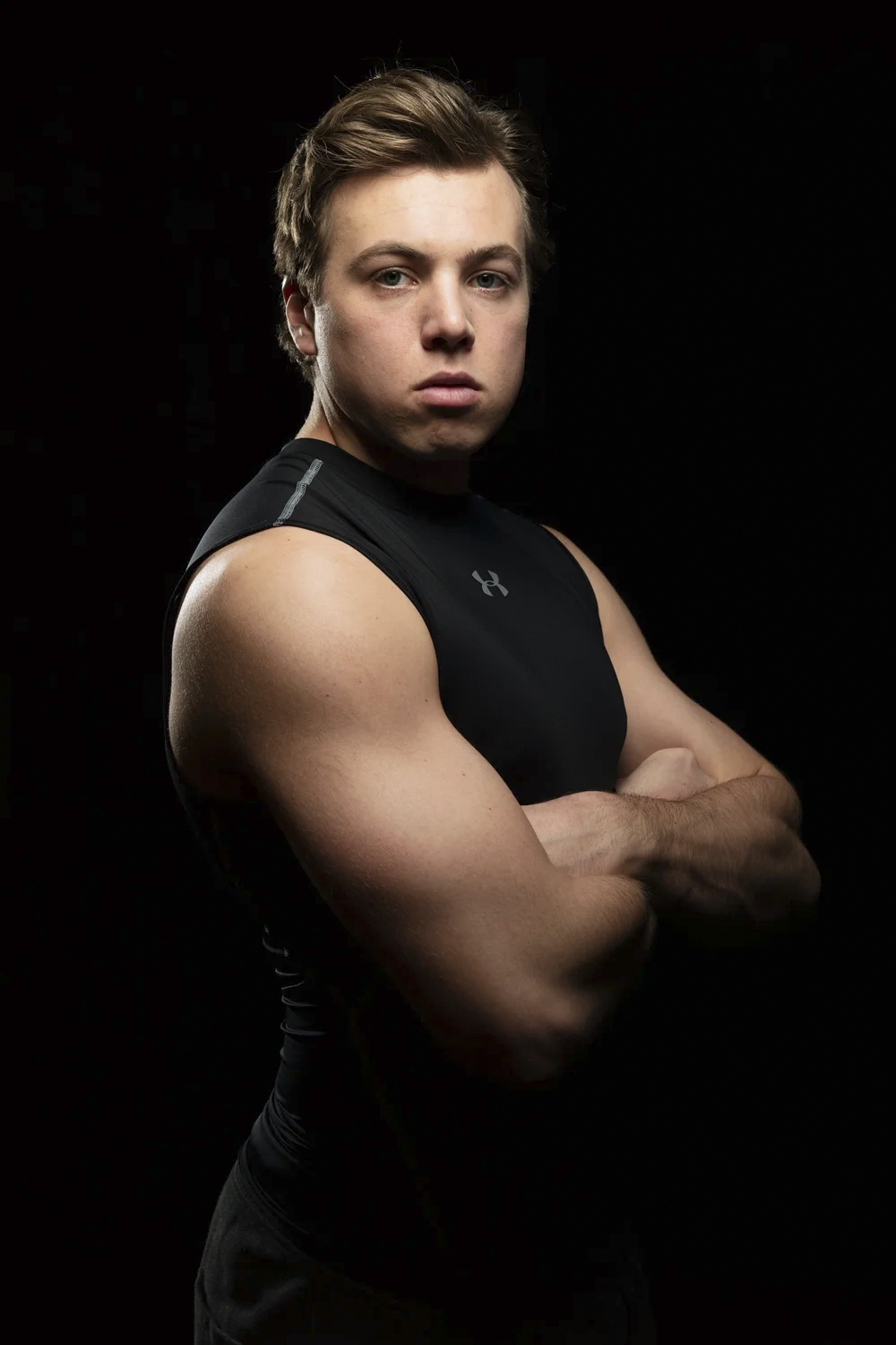 Young man with muscular arms wearing a sleeveless black athletic shirt, crossed arms, against a black background.