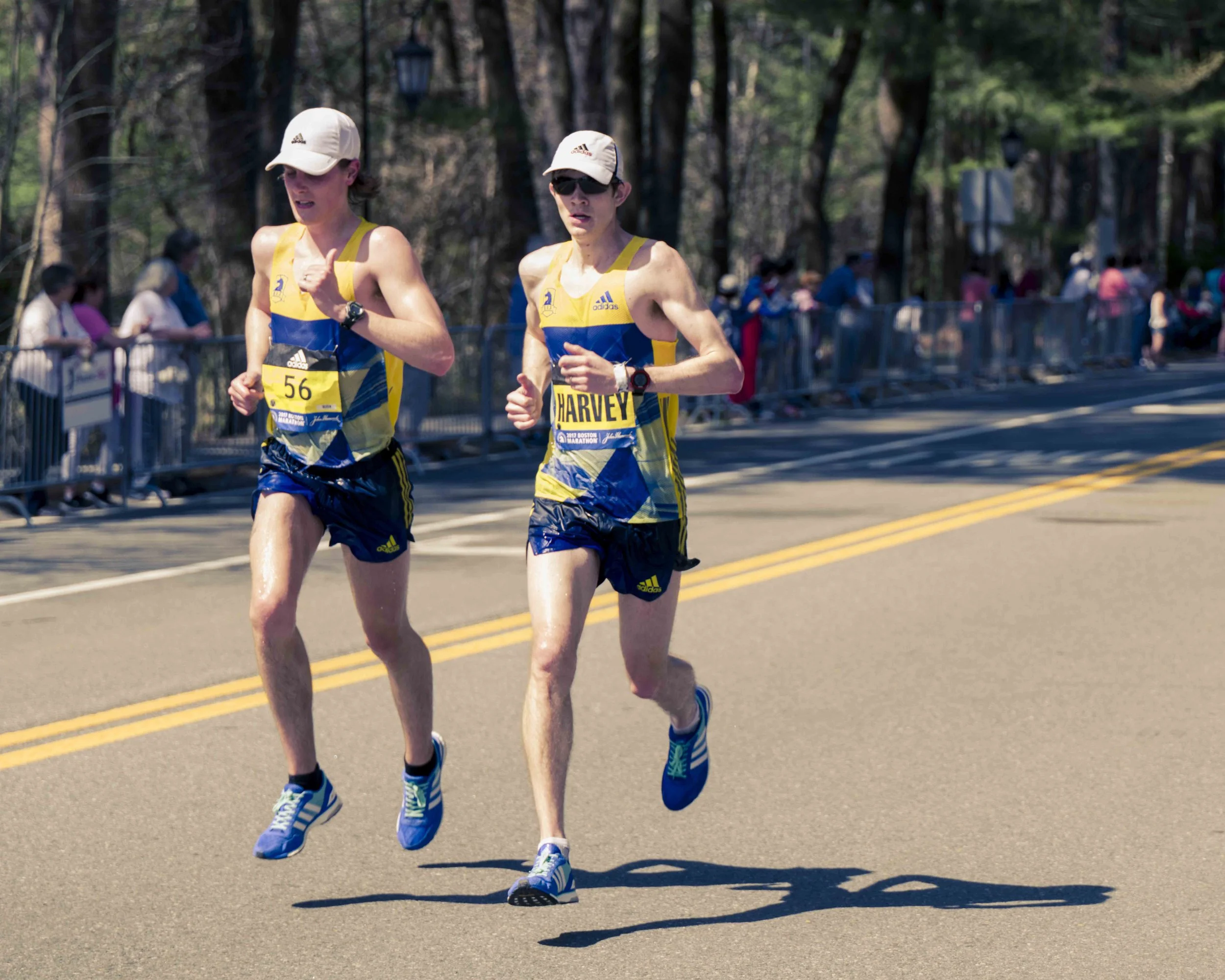 Two male marathon runners wearing yellow and blue athletic gear, hats, and sunglasses running on a street lined with trees and spectators.