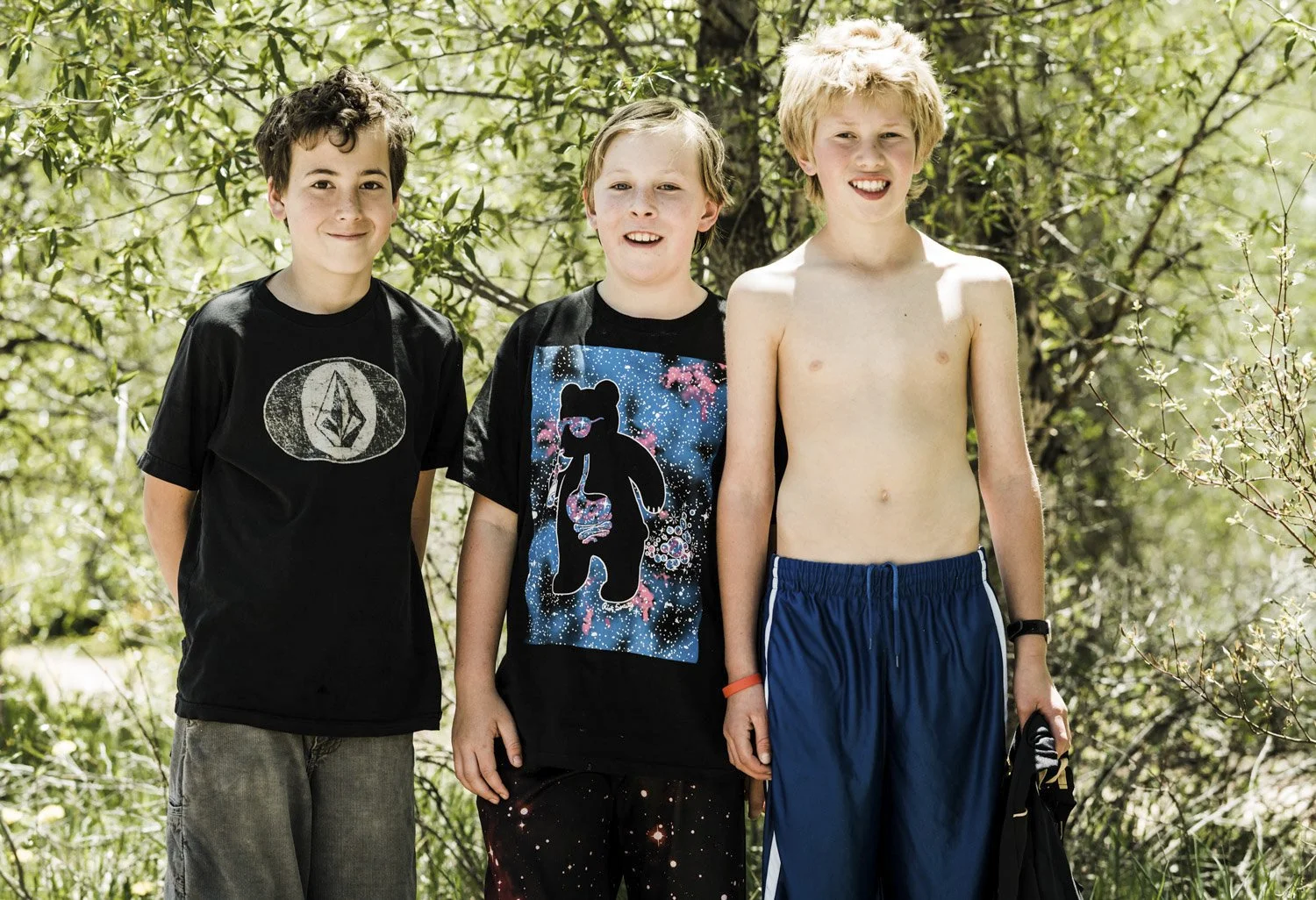 Three boys standing outdoors in front of green trees.