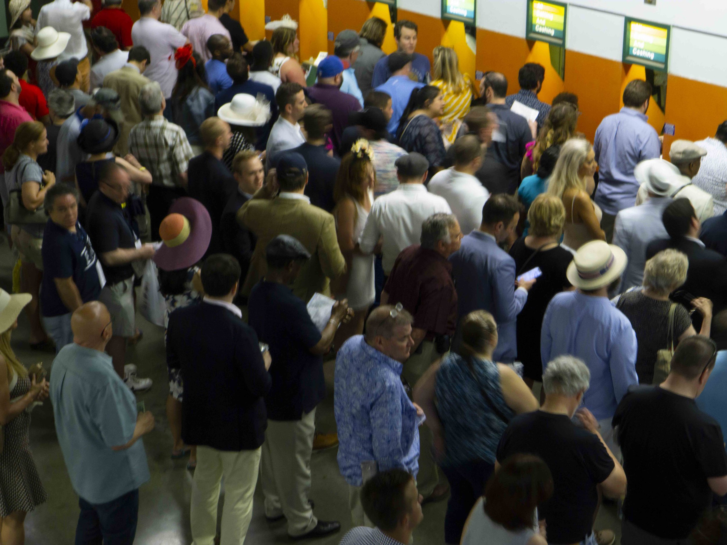 A large crowd of diverse people standing in line at a service counter, possibly at a bank or airport, with some looking at their phones or documents.