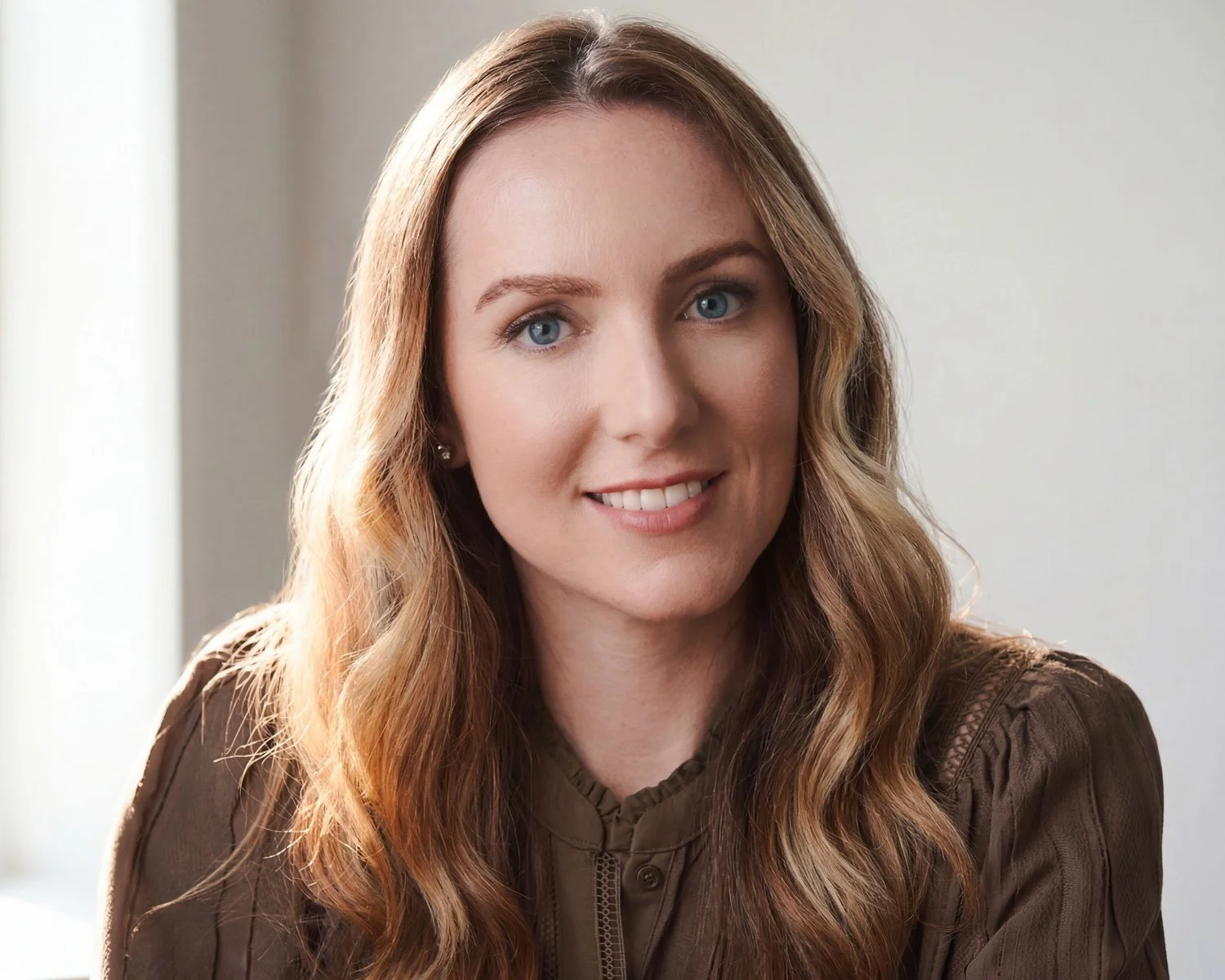 Headshot of a woman with long, wavy blonde hair, blue eyes, and fair skin, wearing a brown top against a soft, medium-tone gradient background.