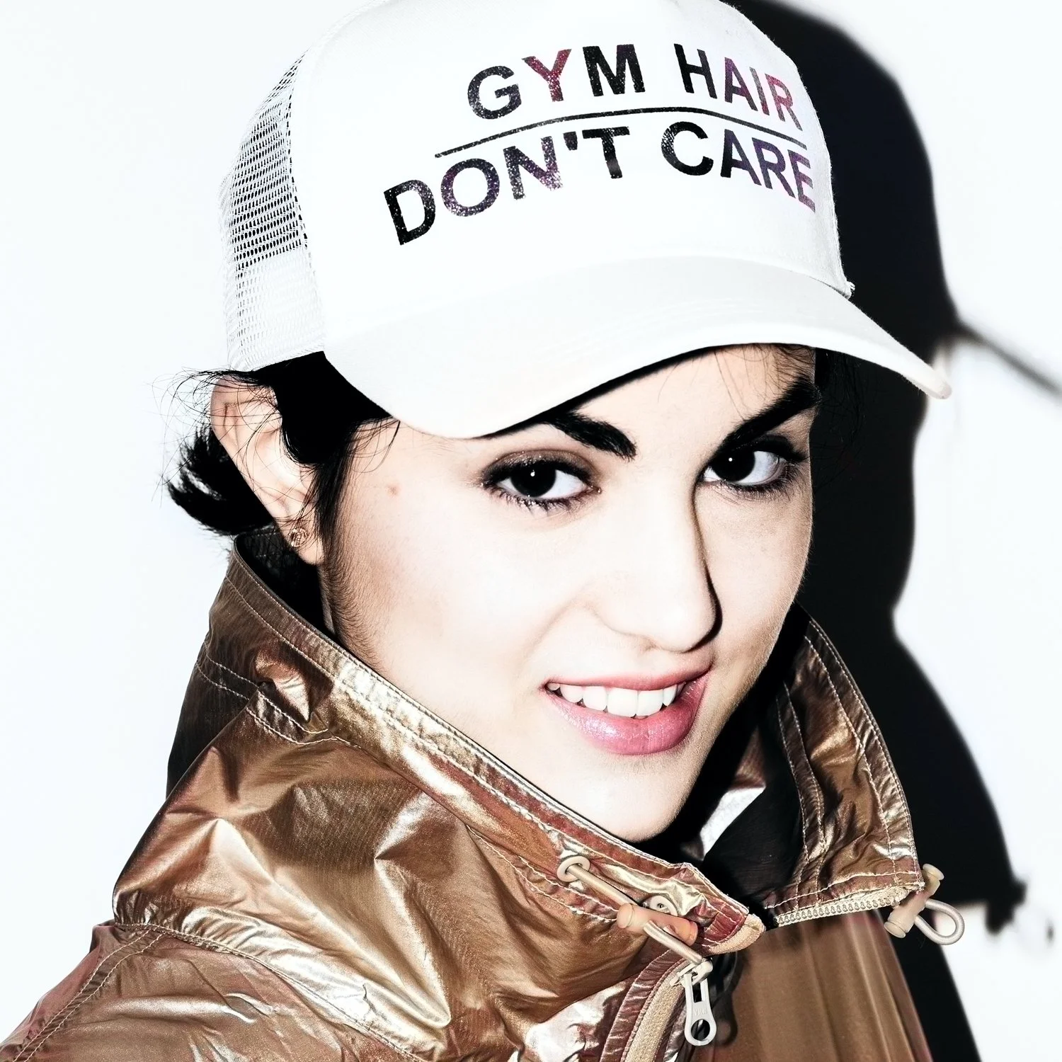 High-contrast flash closeup of smiling young female model against a white background, wearing a gold metallic jacket and a baseball cap with lettering that says Gym Hair Don’t Care.