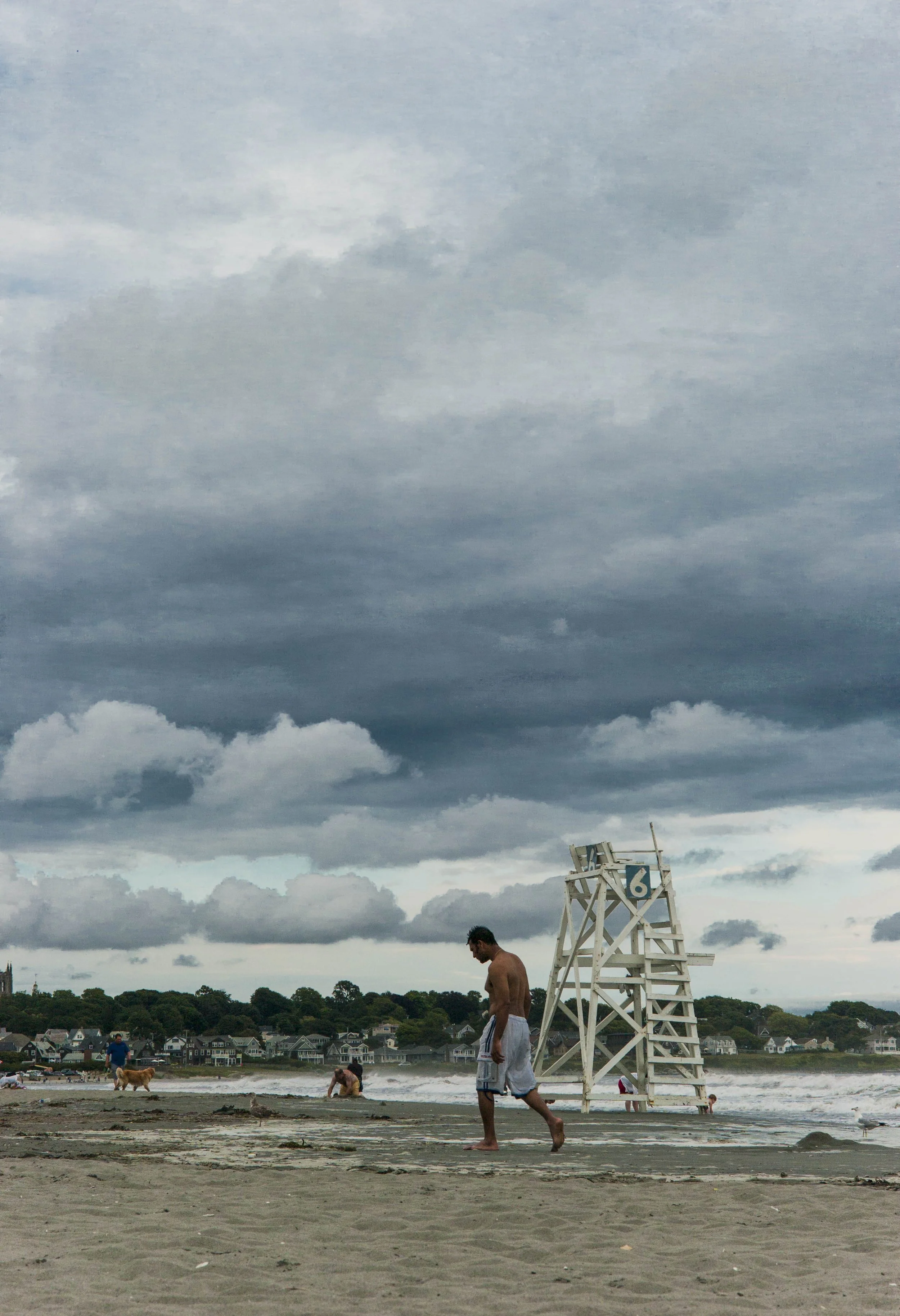 A person walking on a beach with a lifeguard chair marked with the number 6, cloudy sky, and houses in the background.