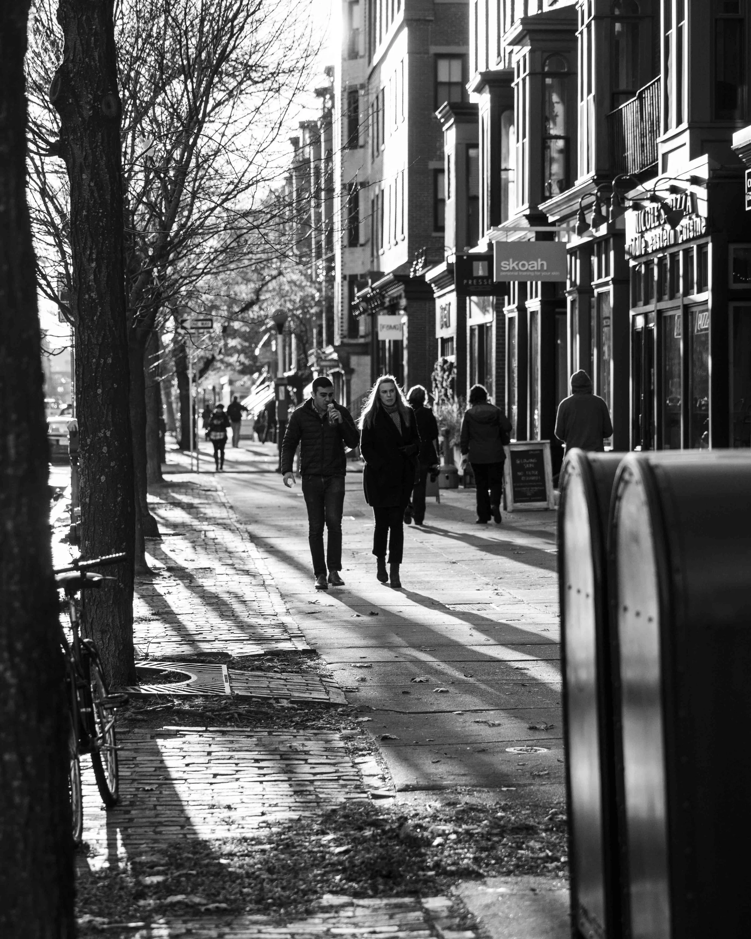 Black and white photo of a city street with pedestrians walking on the sidewalk and storefronts on the right. Trees line the left side of the street, and parked cars are visible in the foreground.