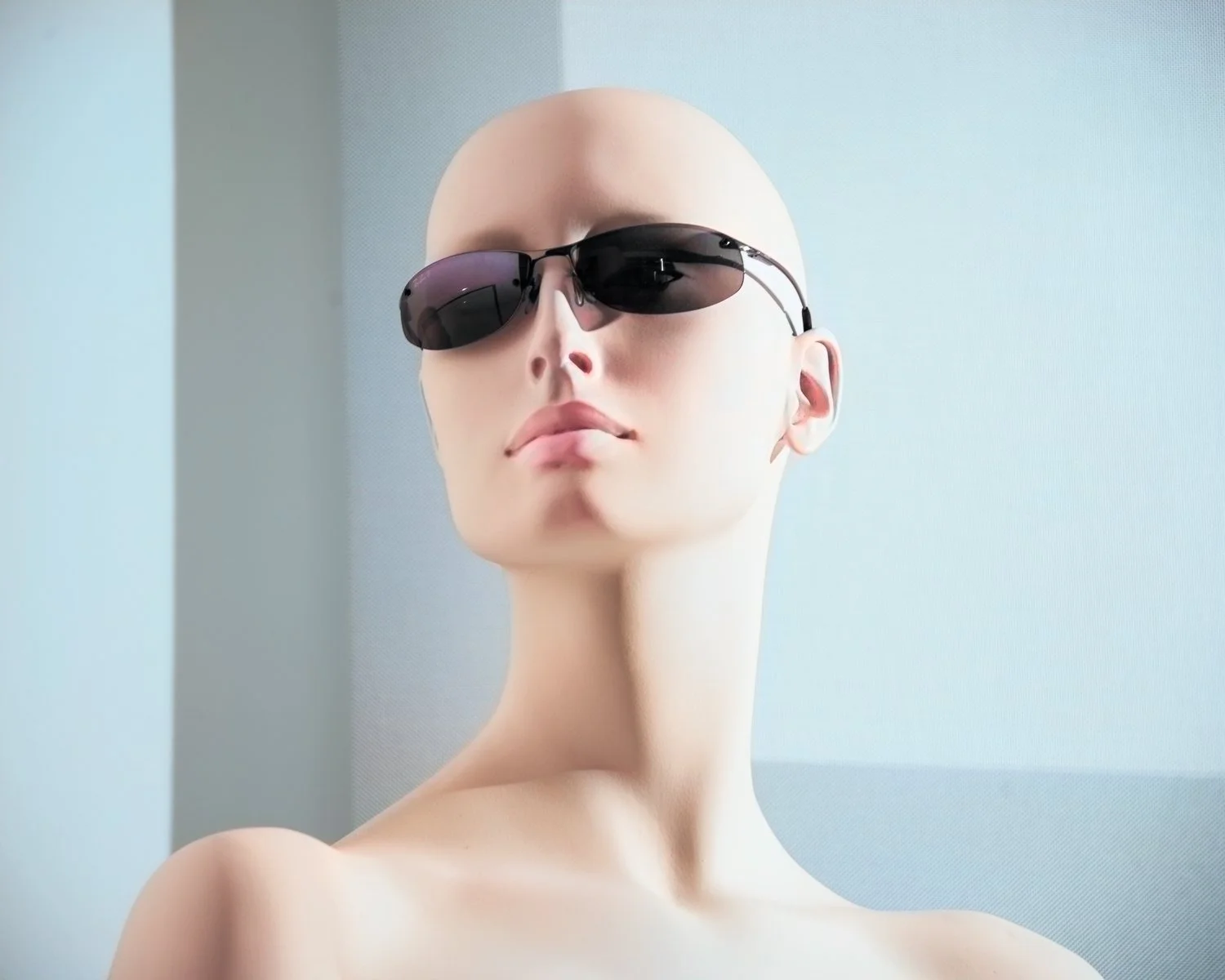 A mannequin head with no hair, wearing dark sunglasses, with a neutral facial expression against a light blue and gray background.