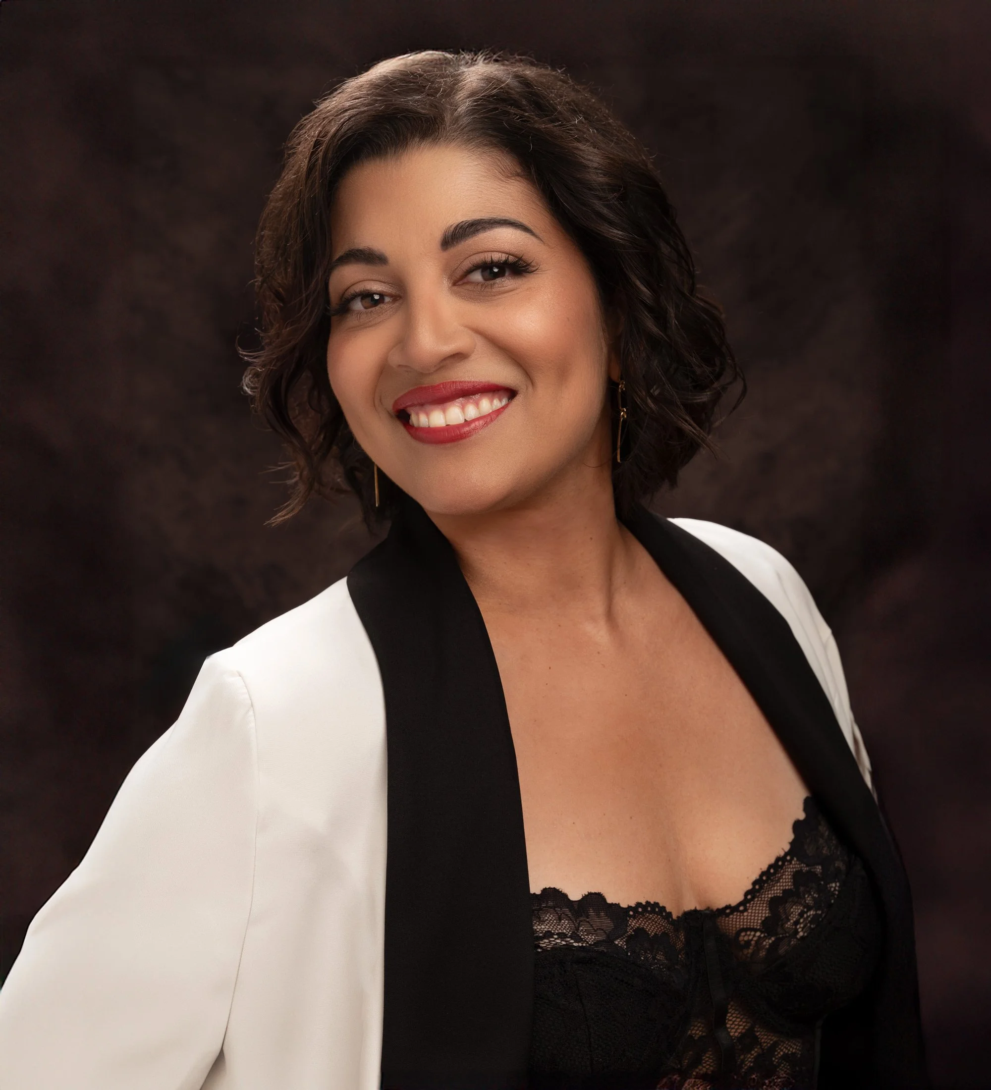 Portrait of smiling, expressive Latina woman with dark hair styled in a bob. She has bright red lips and wears a white and black jacket over a black lace lingerie top. The background is dark brown oil cloth and she is cropped just below the breast.
