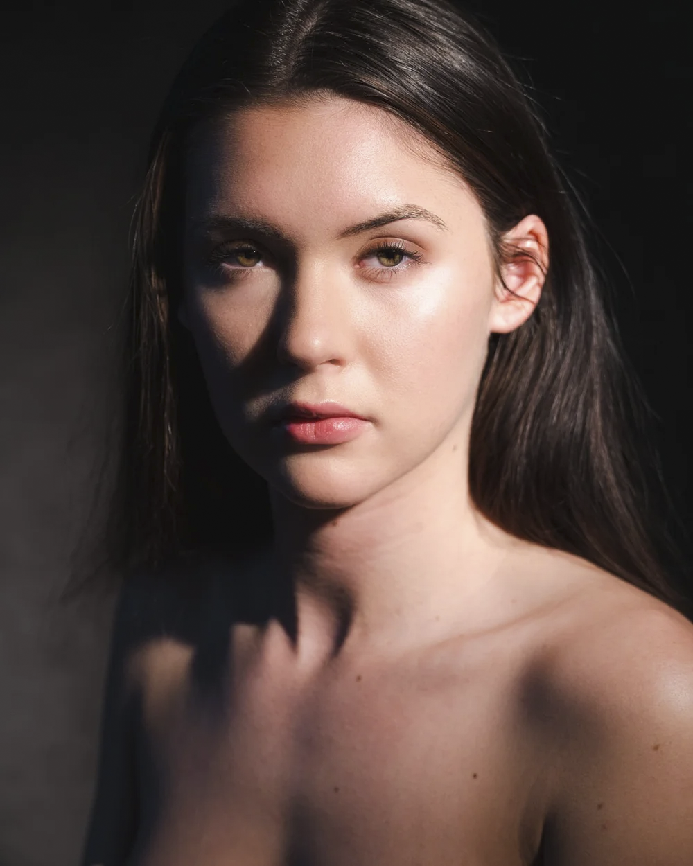Close-up portrait of a young woman with long dark hair, looking directly at the camera with a neutral expression, contrasting lighting highlighting her facial features against a dark background.