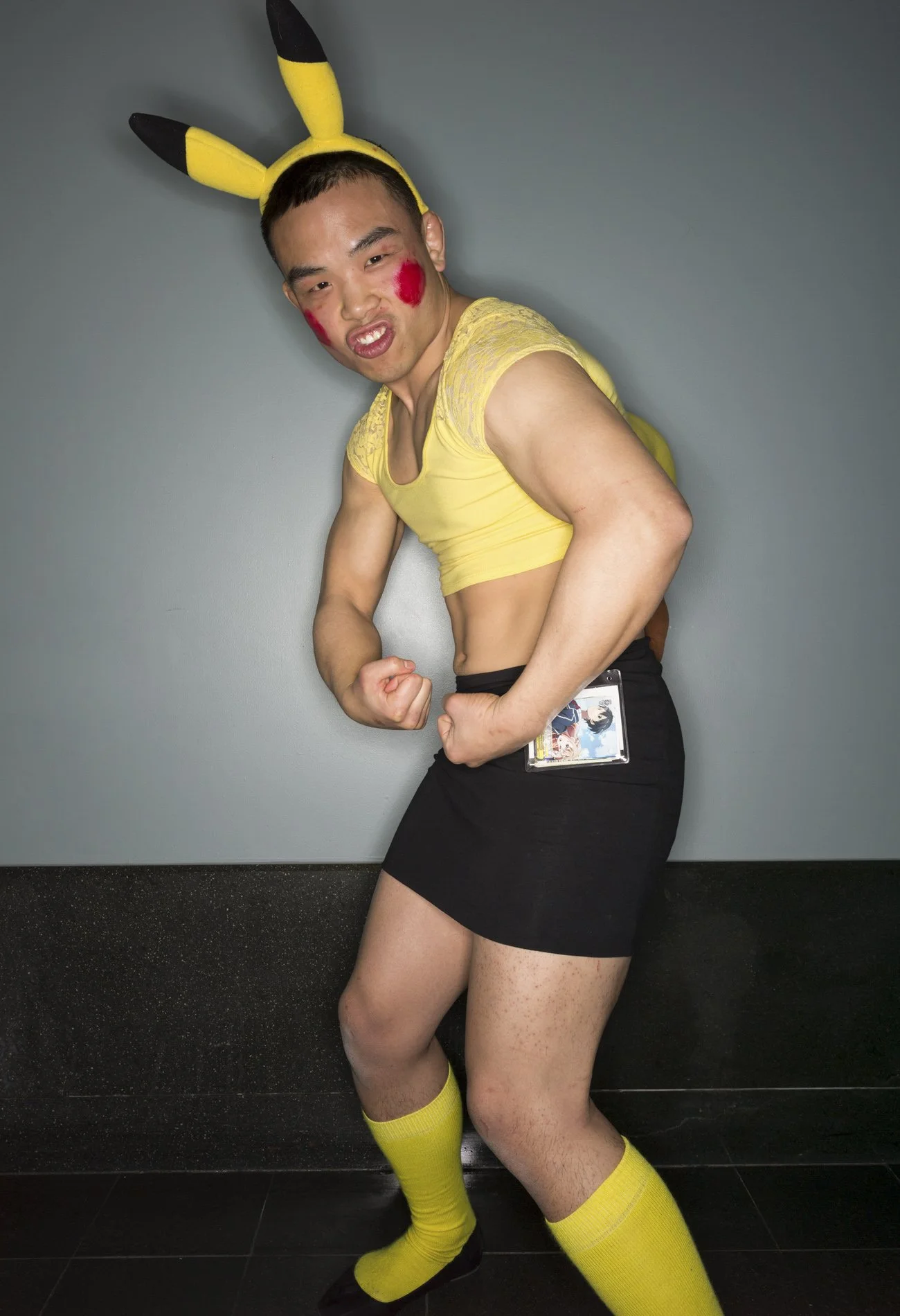 Young man dressed as Pikachu, wearing yellow and black ears, yellow top, black skirt, yellow socks, and black shoes, posing in a playful fighting stance against a gray wall.