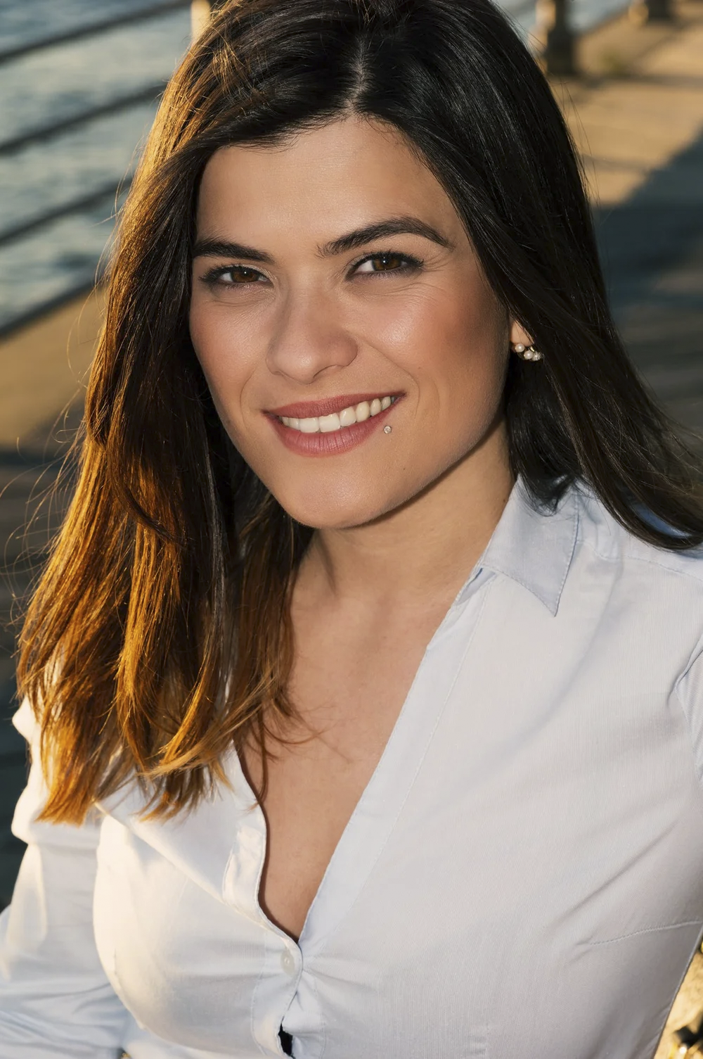A woman with long brown hair, smiling at the camera, wearing a light-colored shirt, standing outdoors near a body of water at sunset.