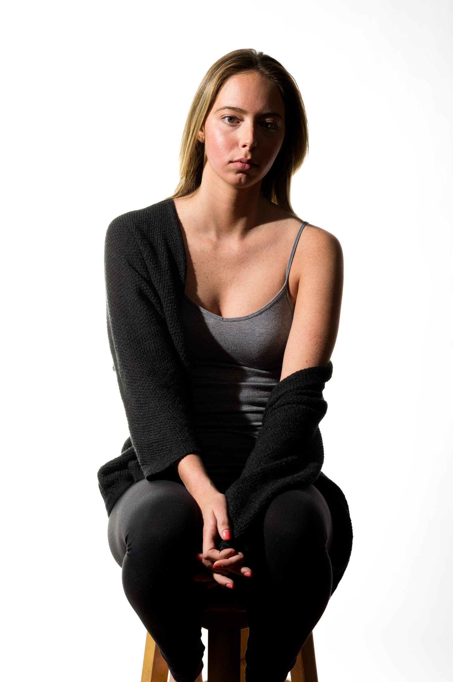 Young female model with long, straight, blonde hair in a gray tank top and black leggings, with a long-sleeve black fleece draped over one shoulder. She sits positioned straight to camera on a wooden stool with hands folded between her legs. The back