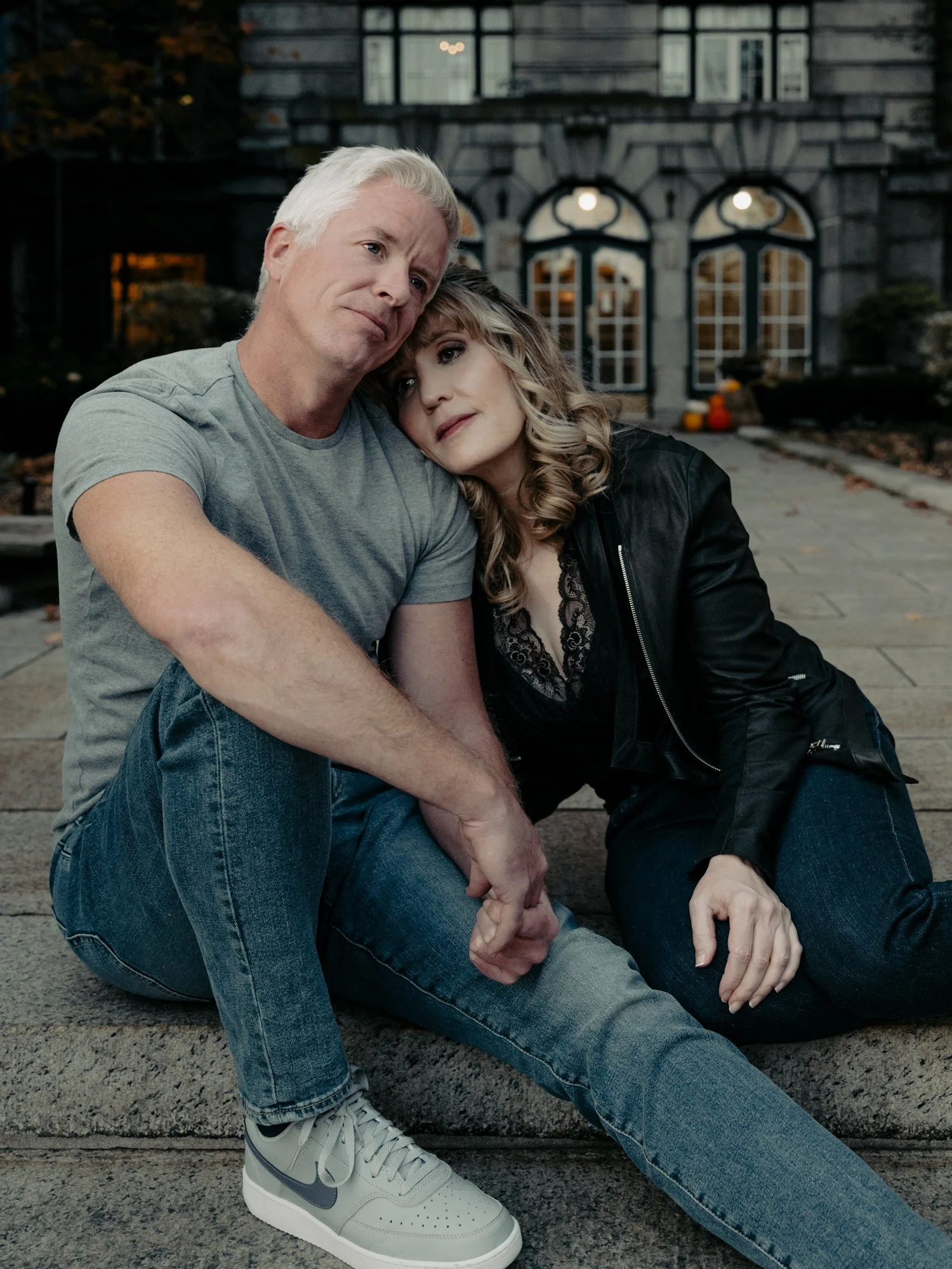A man and a woman sitting on steps outside a building with large windows, holding hands, and leaning on each other. It's evening or late afternoon, and the mood is somber or reflective.