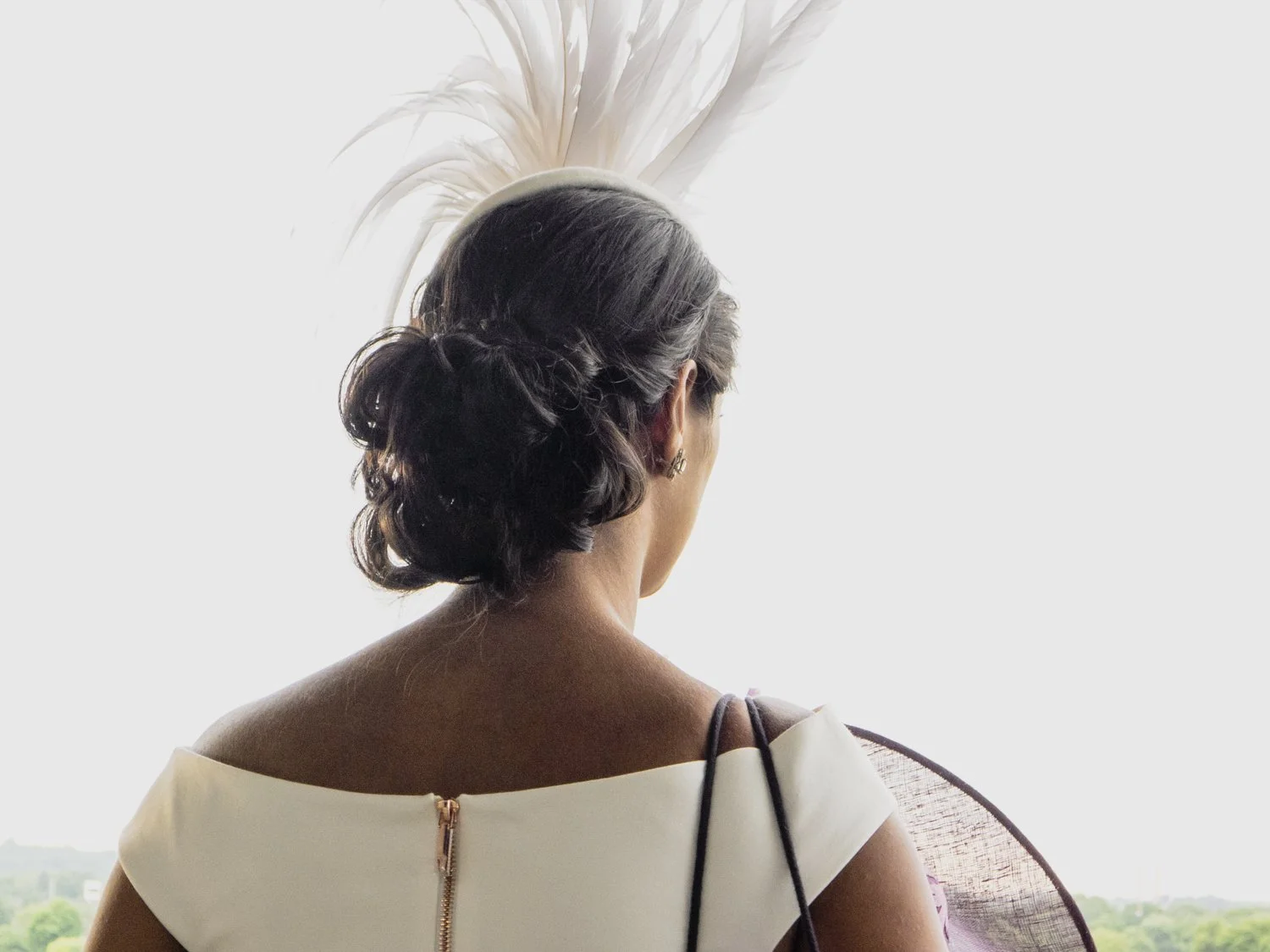 Back view of a woman with dark, curled hair in an updo, wearing a white dress with a visible zipper at the back, looking out a window with a distant landscape.