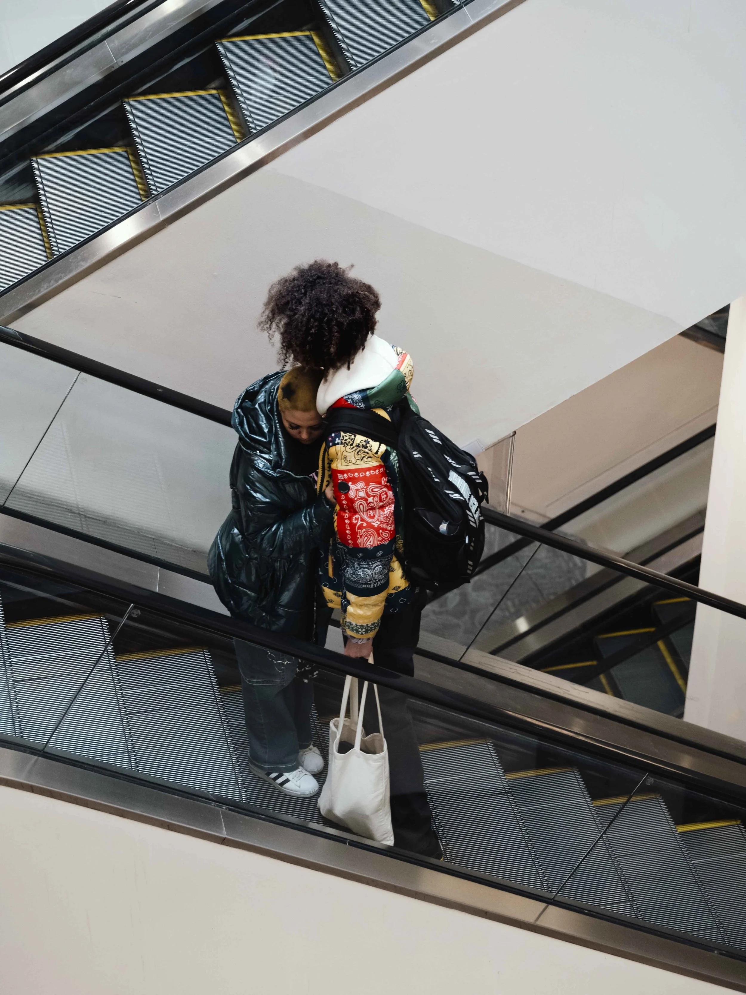 Two women standing on an escalator at an indoor location. One woman with curly hair wearing a shiny black jacket and the other wearing a colorful jacket with many patterns. They are conversing and carrying bags, one with a white tote bag and the othe