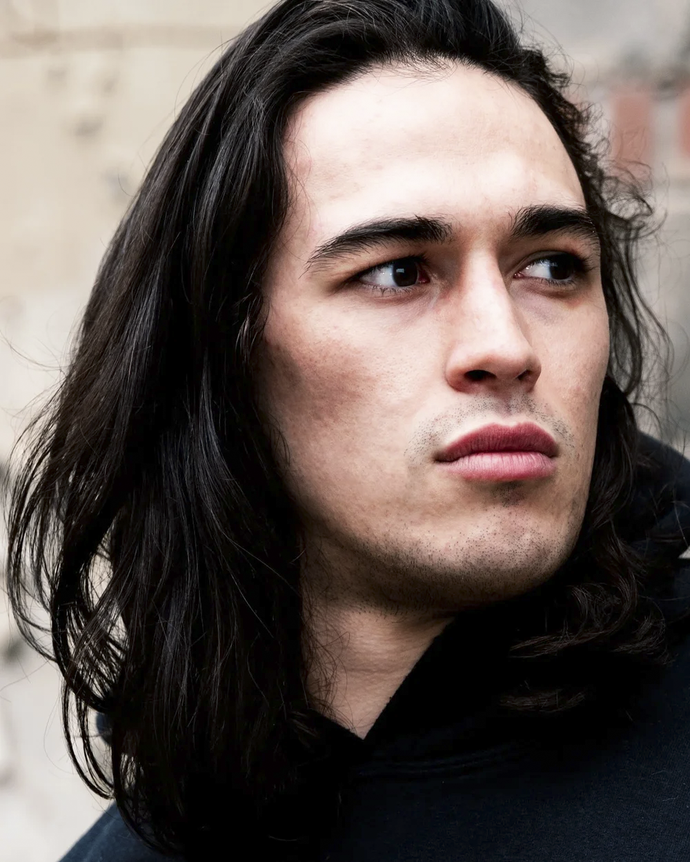 Close-up of a young man with long dark hair and blue eyes, wearing a black jacket, looking thoughtfully to the side, with a blurred brick wall background.