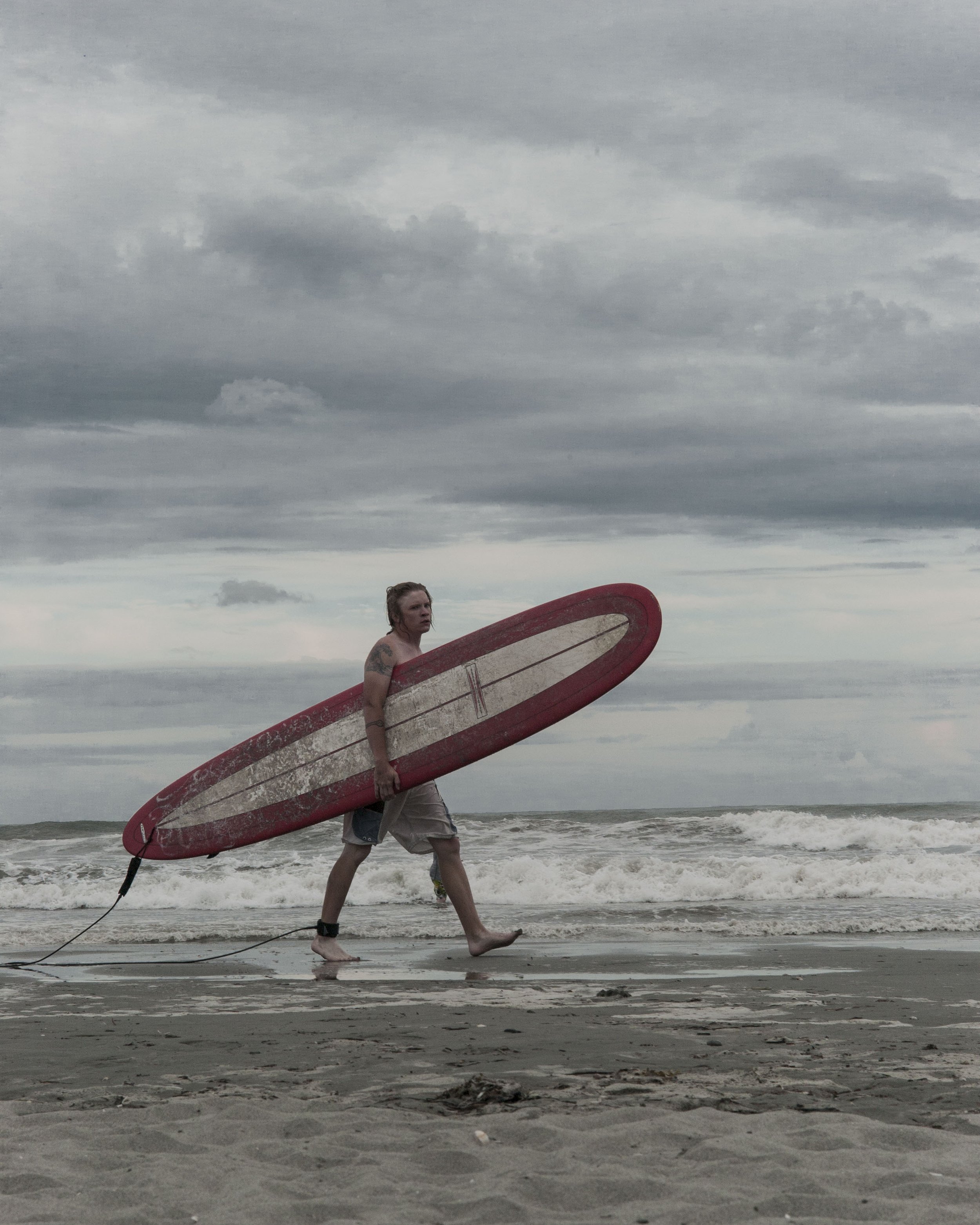 A person with tattoos on their arm and wearing a black ankle strap holding a red and white surfboard walking along a sandy beach near the ocean on a cloudy day.