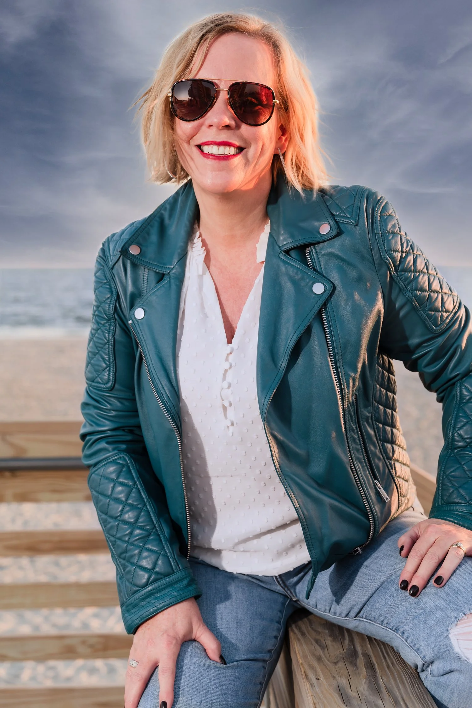 Location portrait of a smiling professional woman in a teal-colored leather jacket with a white shirt and torn blue jeans. Subject has a blonde bob hairstyle and wears red lipstick and dark aviator sunglasses. She sits straddled on a wooden fence wit