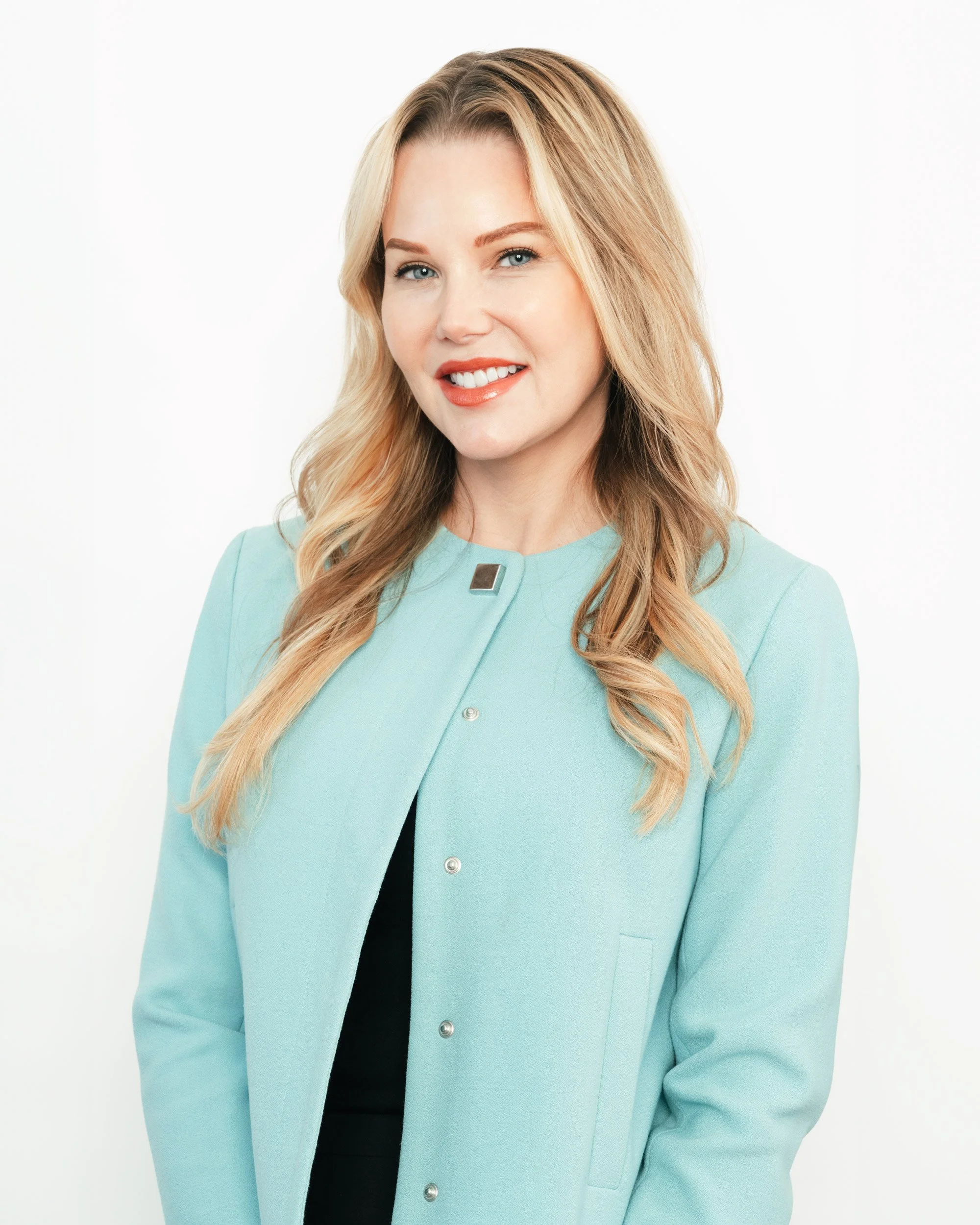 A woman with blonde wavy hair, blue eyes, wearing a mint green blazer over a black top, smiling with open lips, against a white background.