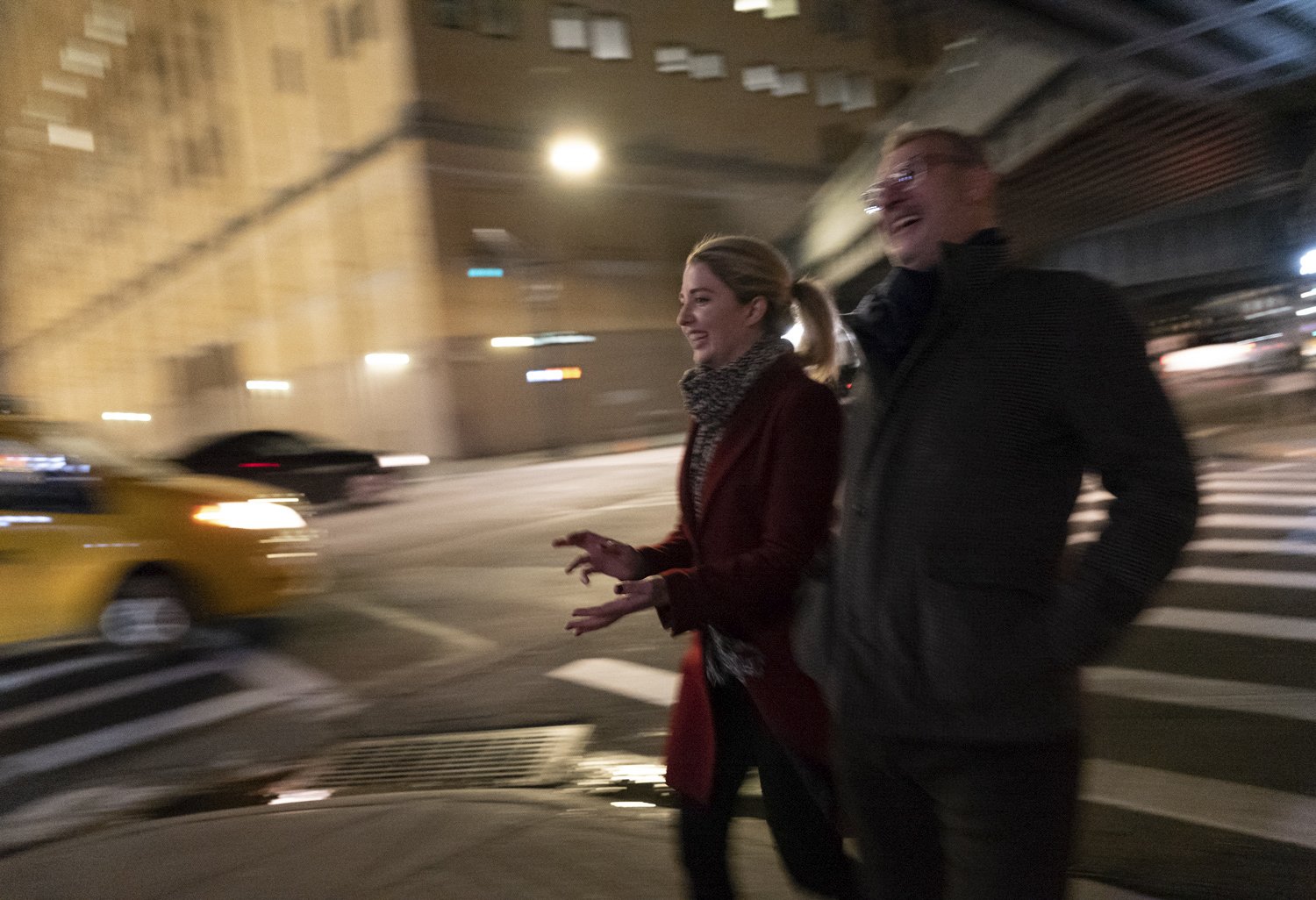 Two people walking across a city street at night, blurred motion of vehicles in the background, city buildings with lights, one woman and one man, smiling and talking.